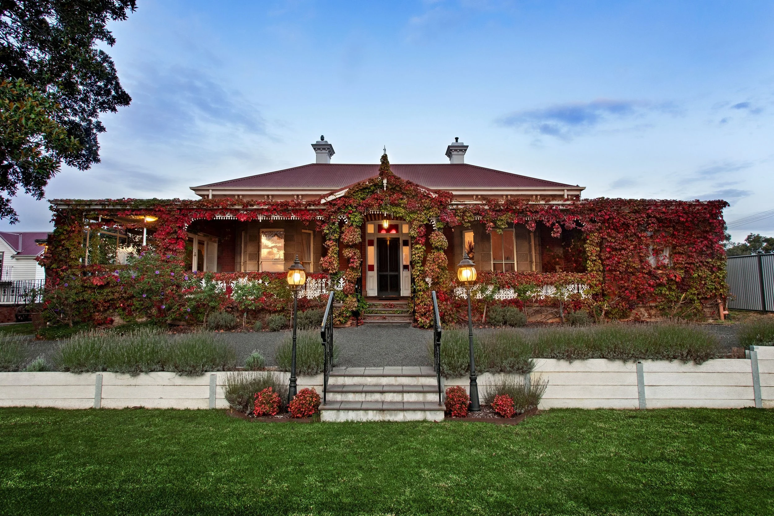 A two-story house with a red roof and brick exterior, covered in red and green ivy, with a front porch and steps, two black lamp posts, and a well-maintained green lawn and flowerbeds.