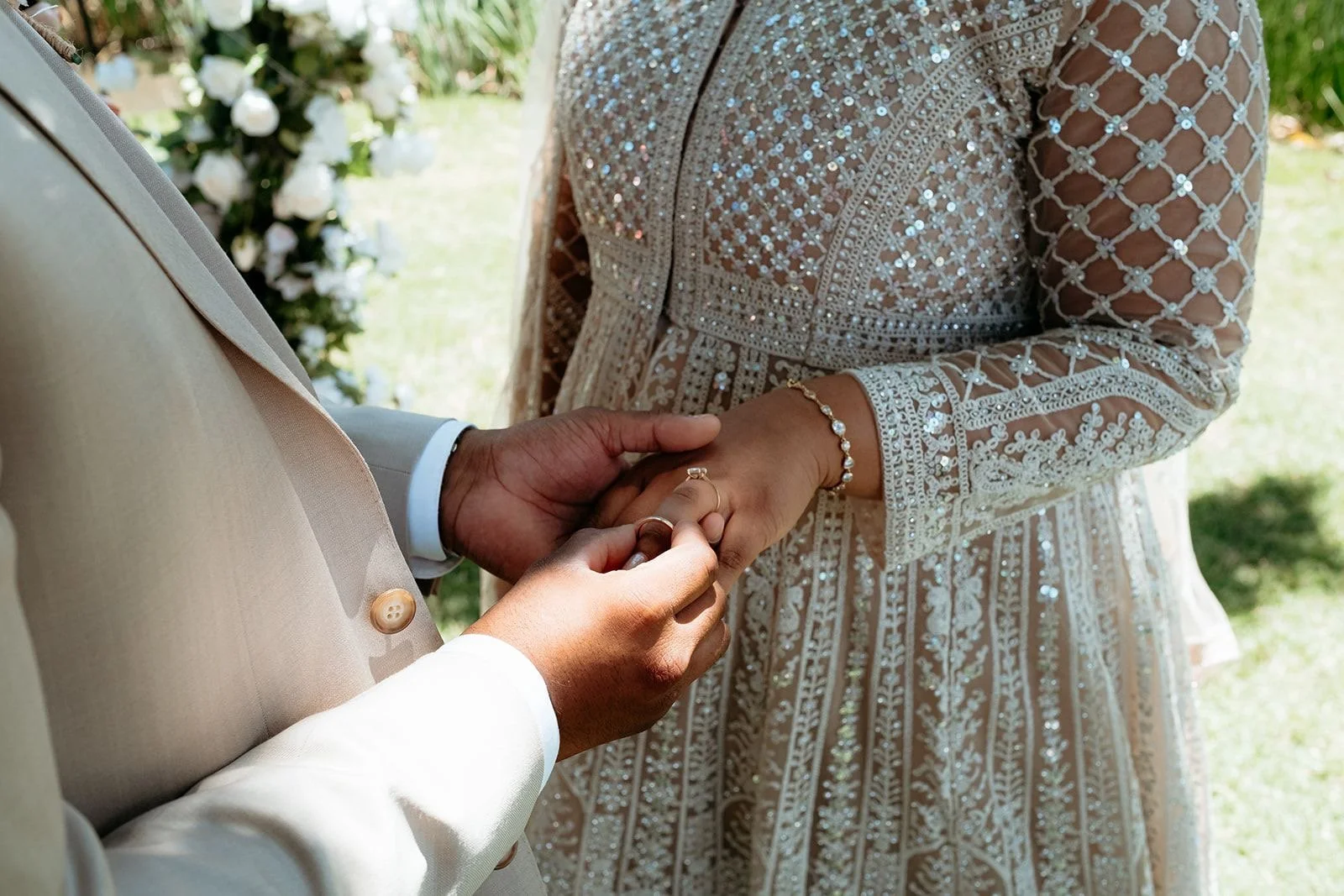 A groom is placing a ring on a bride's finger during a wedding ceremony outdoors. The bride wears a detailed, beaded lace dress and jewelry, and the groom is in a light-colored suit.