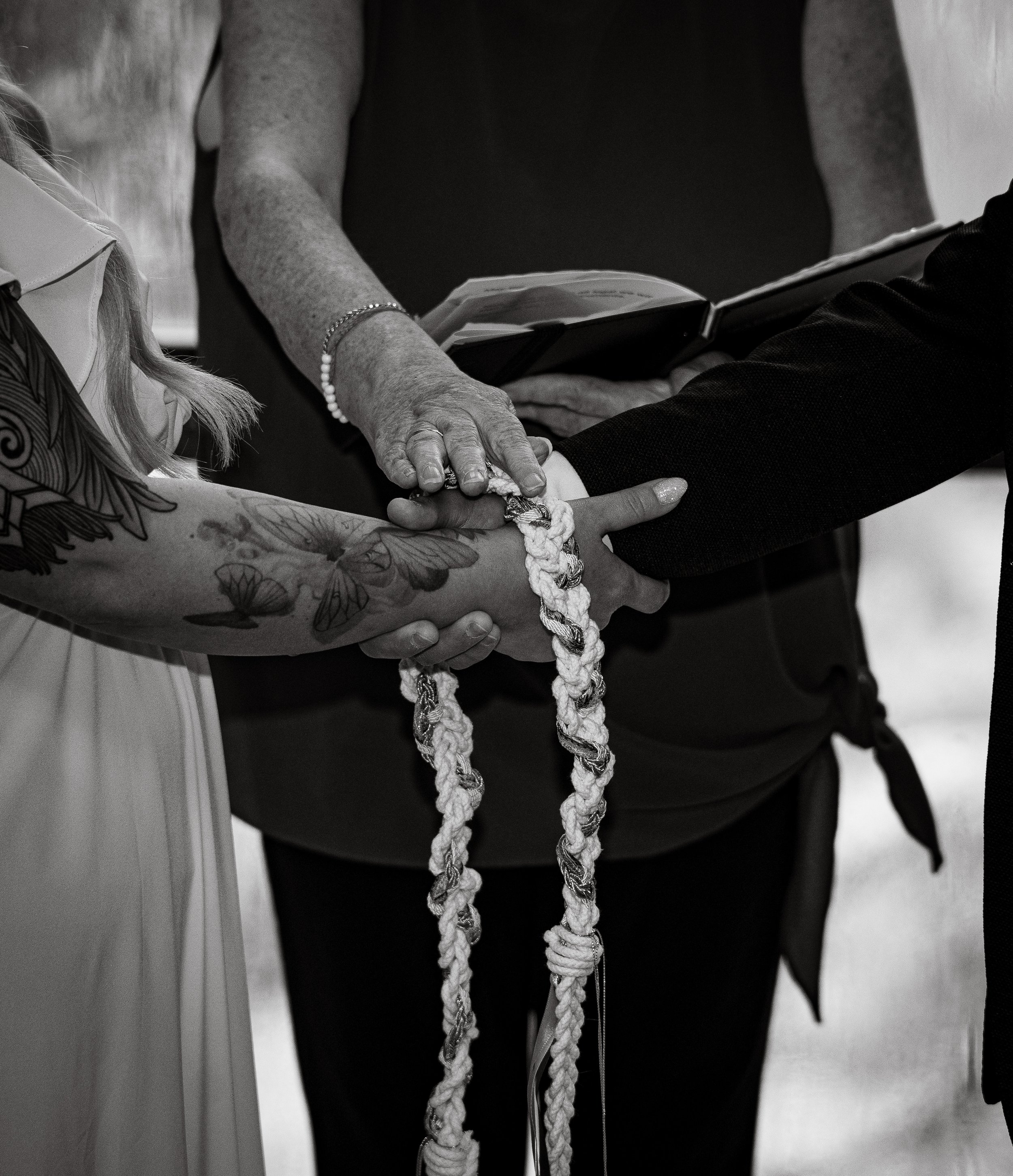 A black and white photo of a wedding ceremony, showing two people holding hands with a officiant. The person on the left has tattoos on their arm, and both hands are linked with a decorative rope or cord. The officiant is holding a book, and the focus is on the hands and the ceremonial cord.