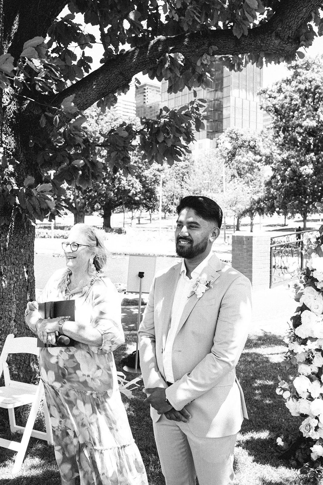 A man dressed in a light suit with a boutonniere stands outdoors under a tree, smiling, during a wedding ceremony, with a woman holding a book and microphone nearby, in a park setting with trees and buildings in the background.