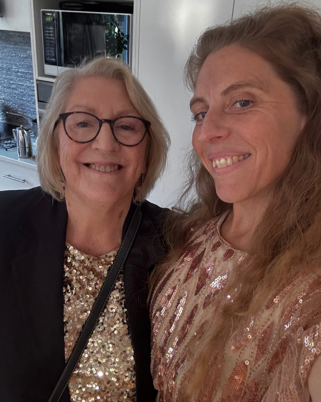 Two women smiling in a close-up selfie, one with glasses and the other with long wavy hair, in a kitchen setting with a microwave in the background.