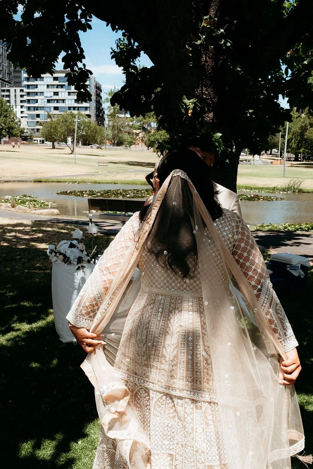 A bride in a traditional, intricate lace and bead wedding dress holding the edge of her veil outdoors under a tree, with a scenic park and pond in the background.