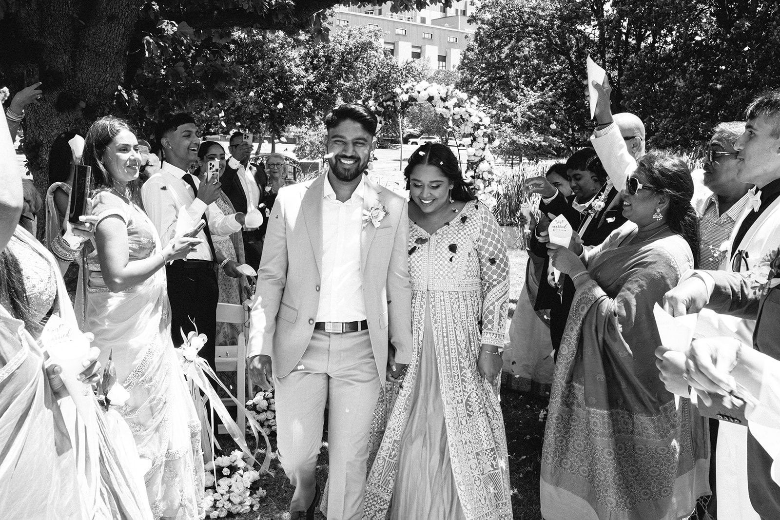 A black and white photograph of a wedding scene showing a smiling couple walking hand-in-hand through a gathering of friends and family outdoors. The groom is wearing a light-colored suit and the bride is dressed in a traditional outfit. Guests are c
