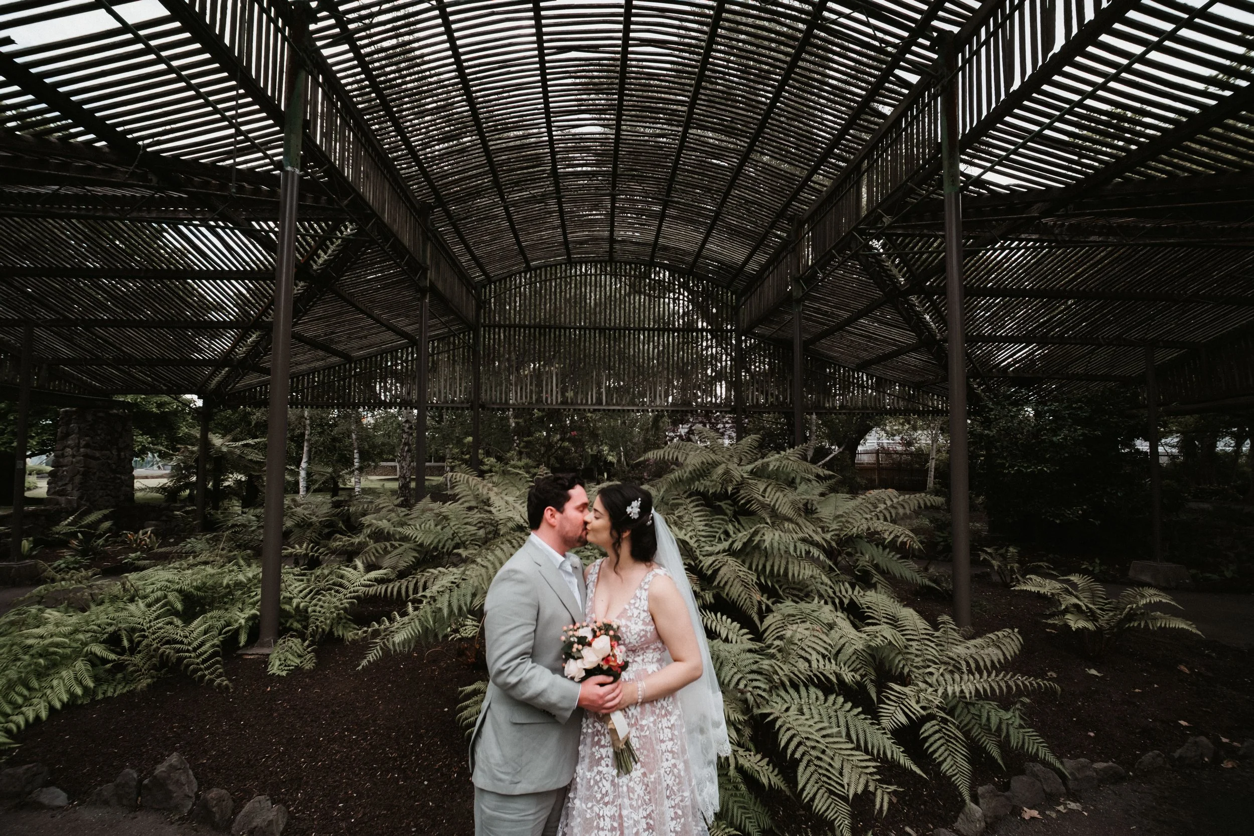 Couple getting married, kissing, standing in a garden with lush ferns, under a large metal structure with a curved ceiling.