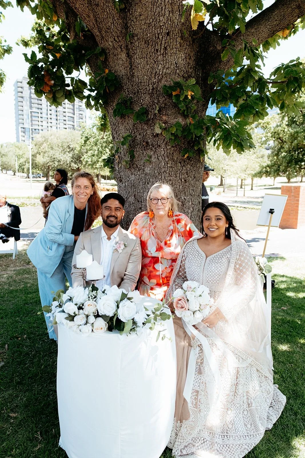 A group of five people, including a bride holding a bouquet, posing outdoors under a large tree at a wedding celebration. The group includes a woman in a light blue suit, a man in a beige suit, an older woman in a colorful blouse, and a woman in a wh