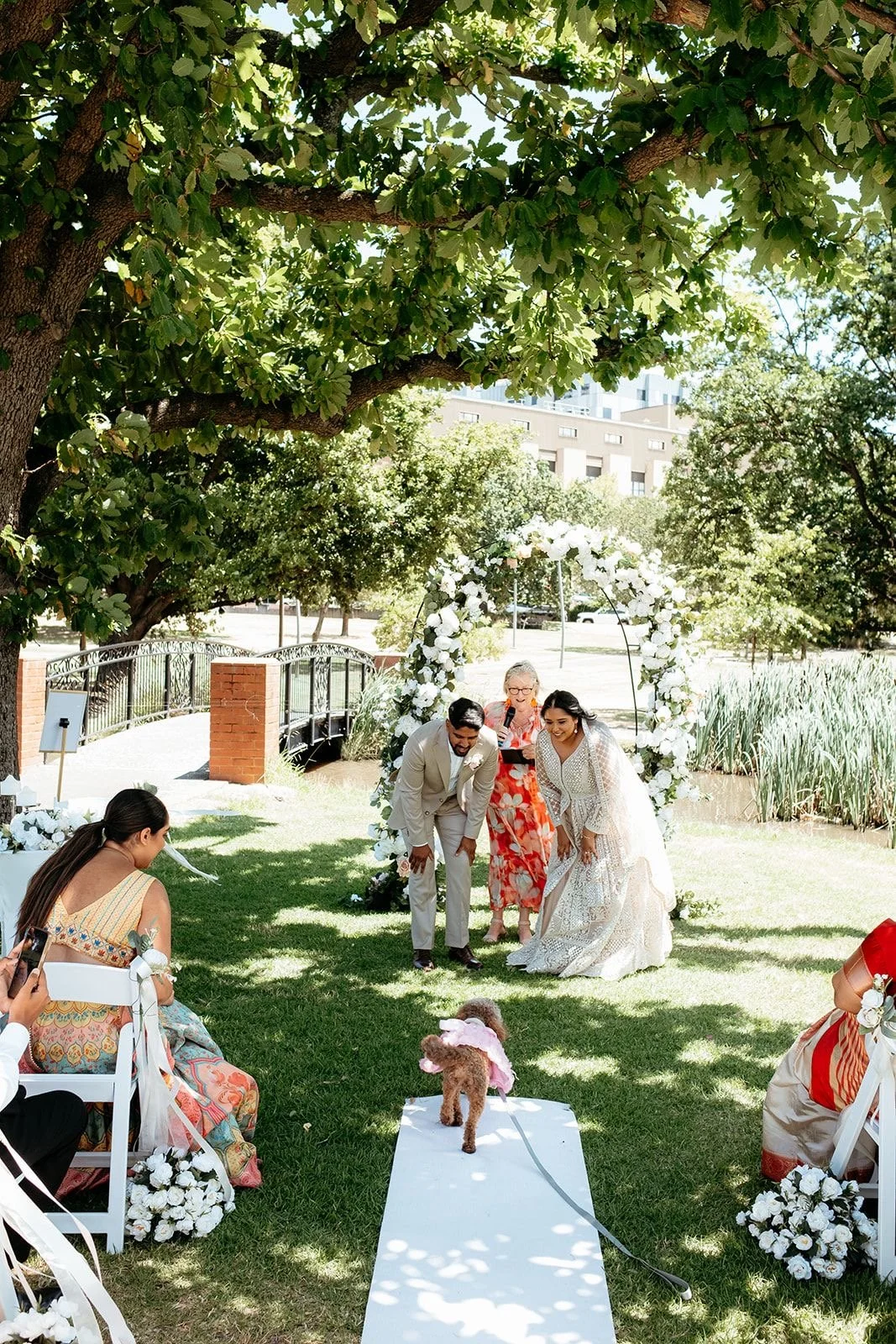 Couple getting married outdoors under a floral arch with guests seated nearby, and a small dog walking on a white aisle.