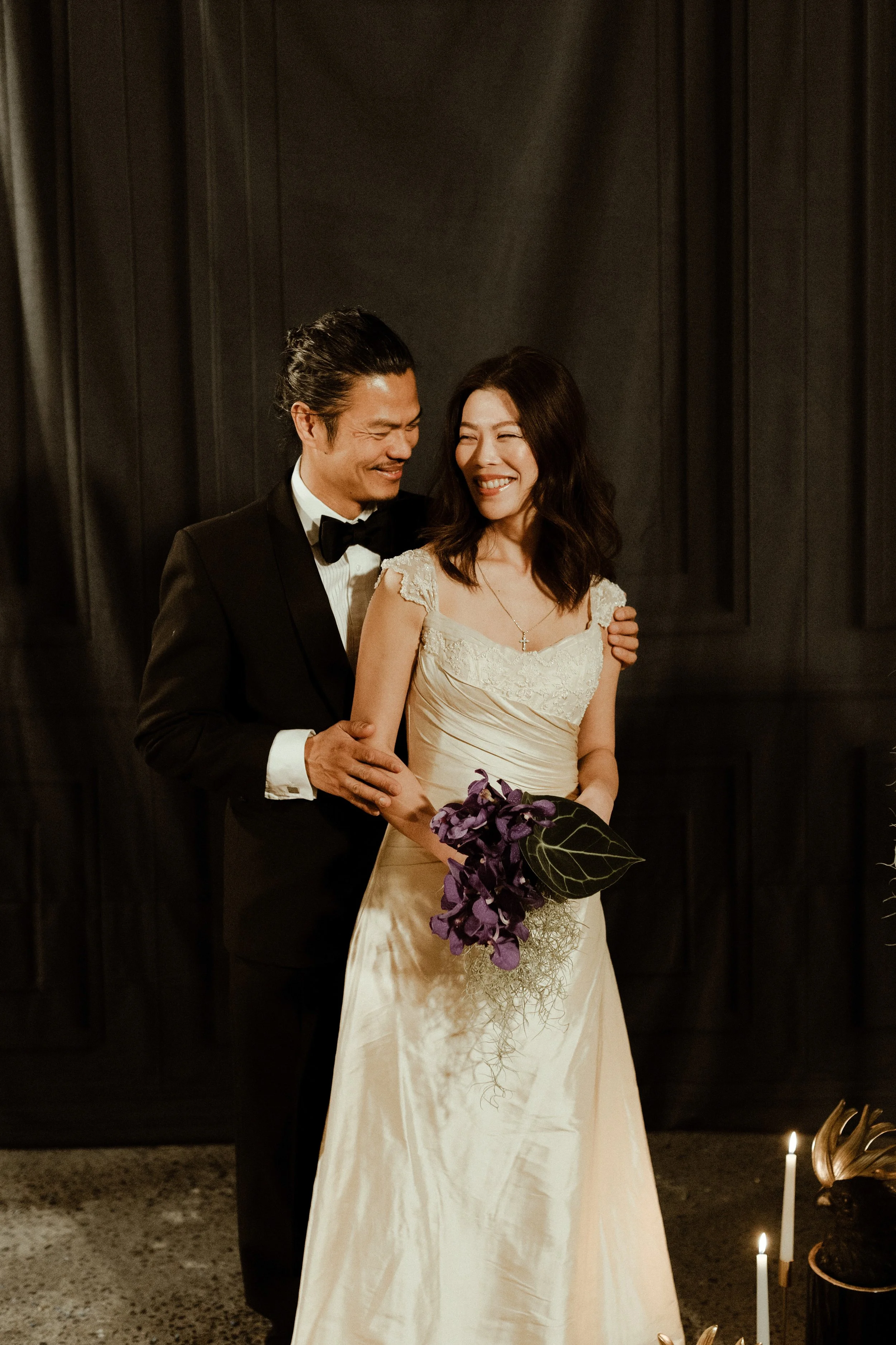 A happy couple in wedding attire, a man in a black tuxedo and a woman in a cream wedding dress, smiling and embracing, with the woman holding a purple flower bouquet, standing in front of a dark background with candles.
