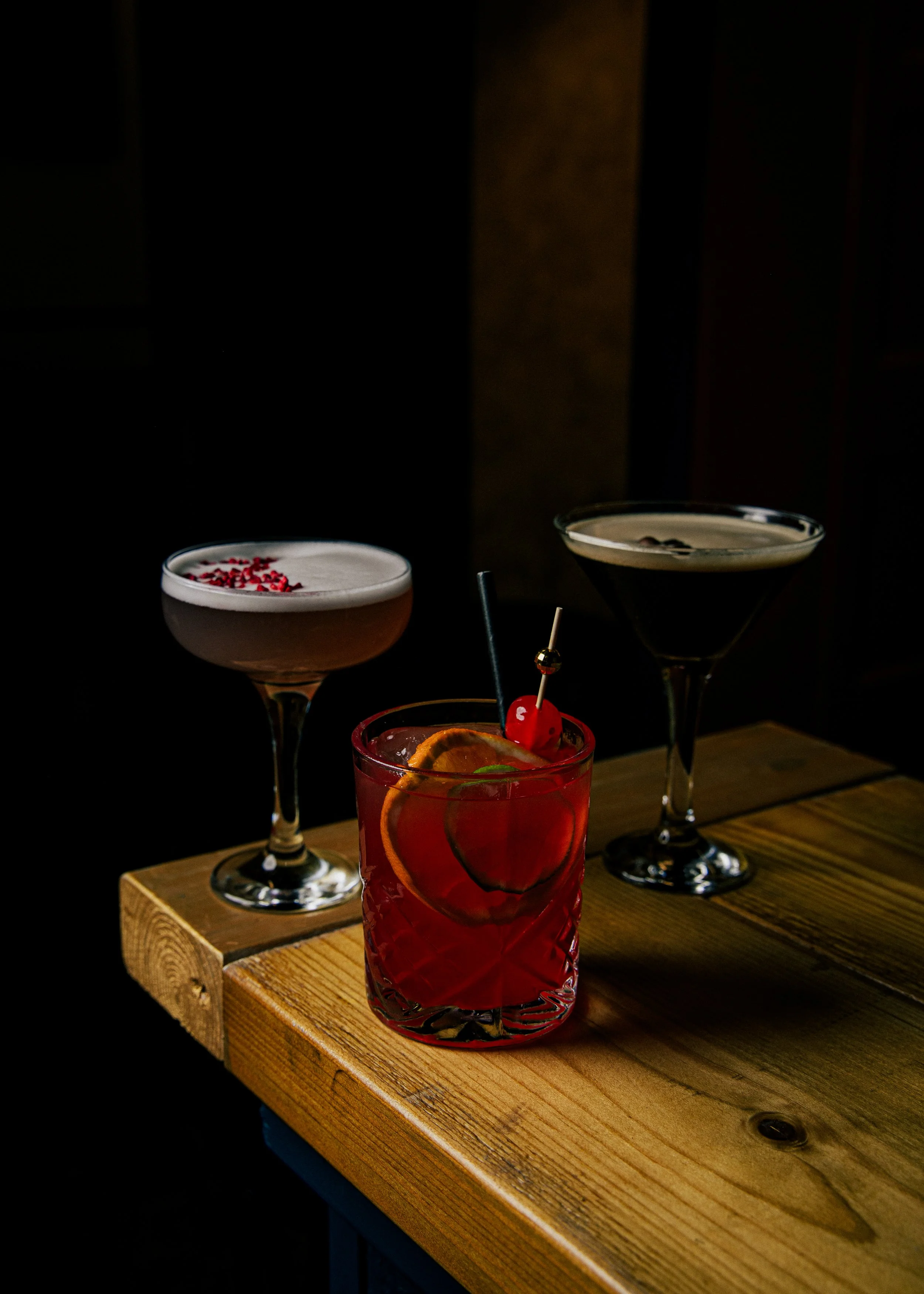 Three cocktails on a wooden table in a dark setting, with one in a short glass garnished with an orange slice, a cherry, and a straw, and two in martini glasses, one with pink foam and red sprinkles, and the other with a clear liquid and a dark garni