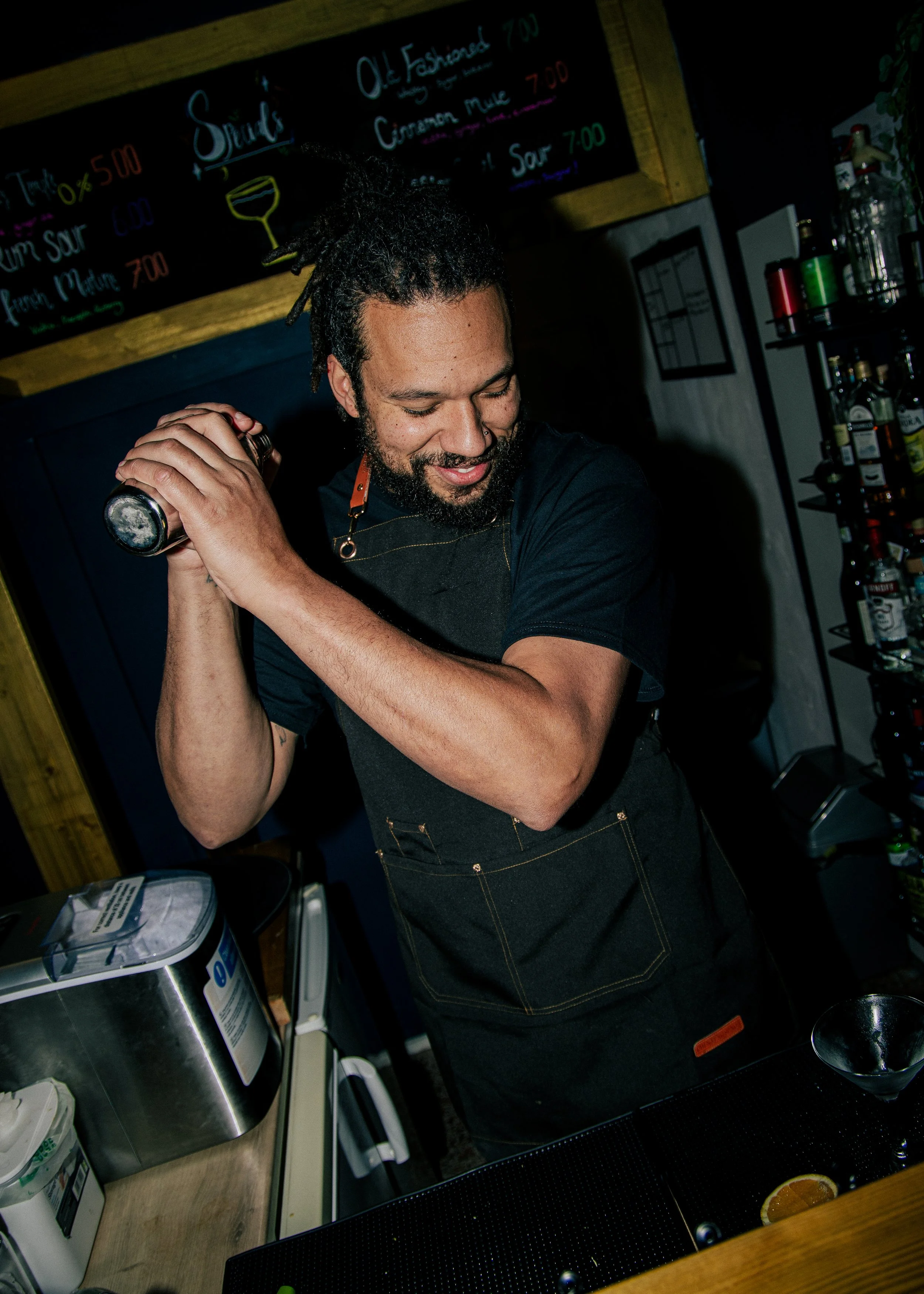 A smiling bartender with dreadlocks in a black shirt and apron shaking a cocktail shaker behind the bar.