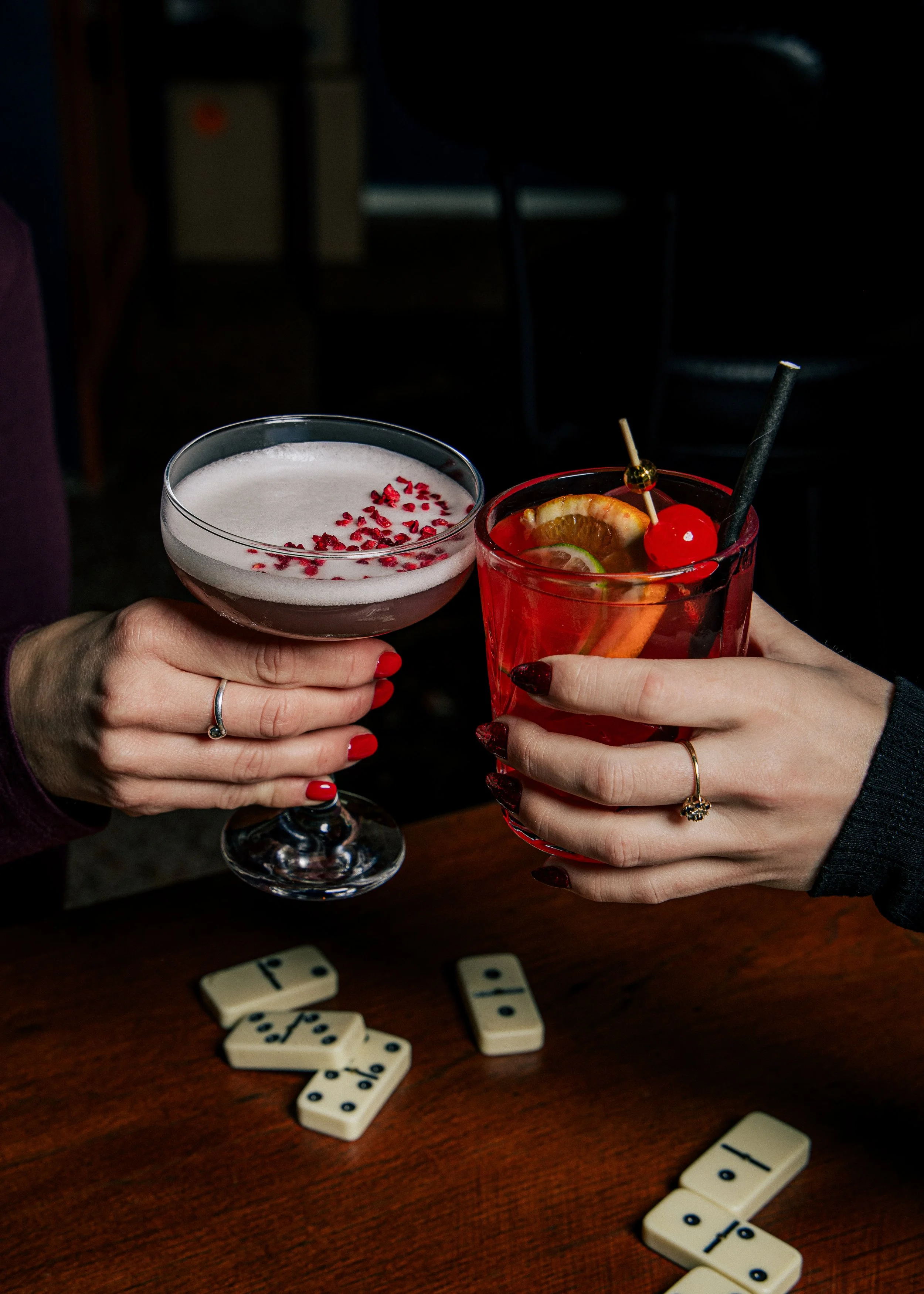 Two people clinking glasses, one with a red cocktail garnished with lemon slices and cherries, and the other with a creamy drink topped with red sprinkles, on a wooden table with domino tiles.