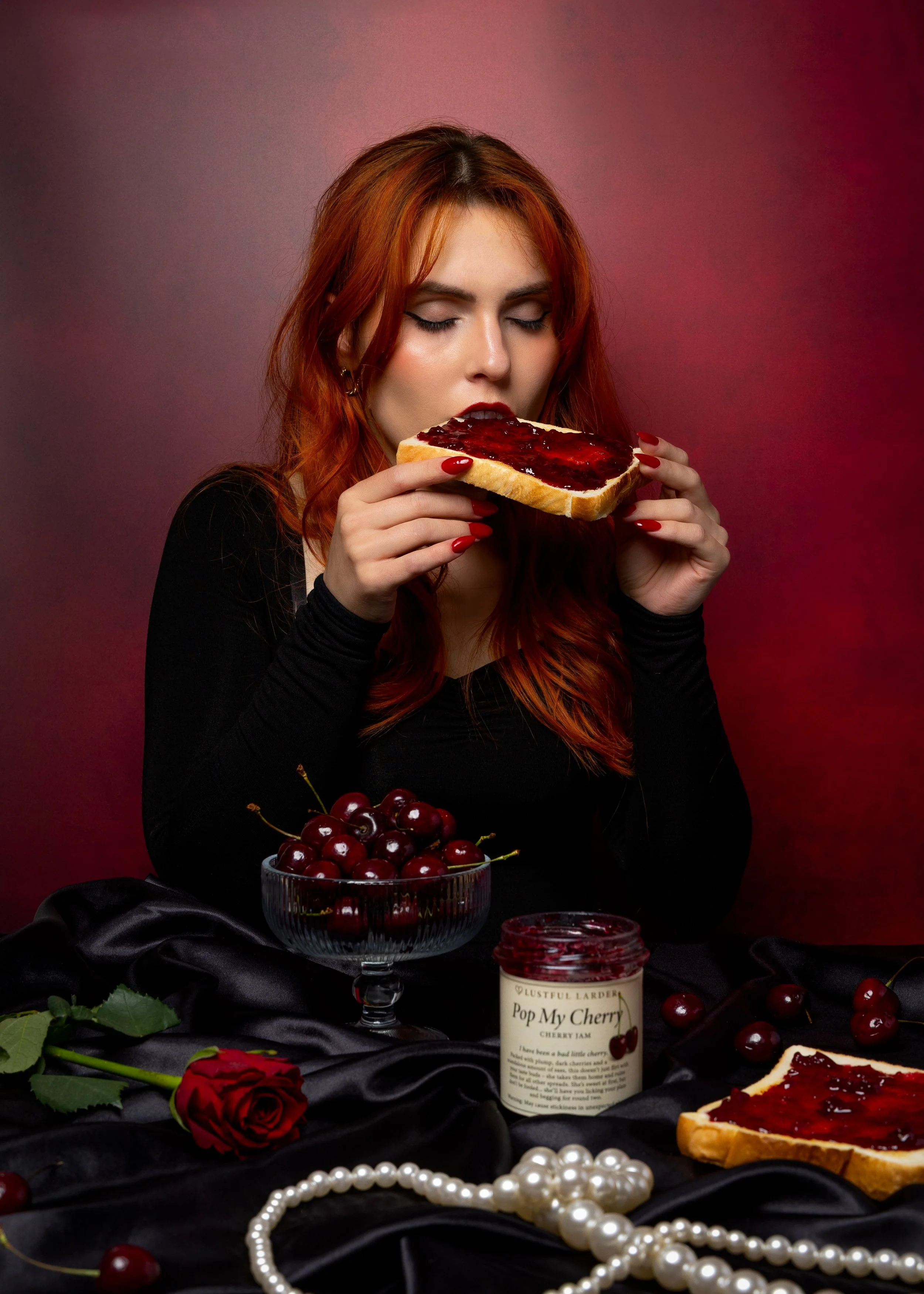 A woman with red hair and black clothing is holding a slice of toast with cherry jam. There is a bowl of cherries, a jar of cherry jam labeled 'Pop My Cherry', a red rose, and a pearl necklace on a black satin cloth in front of her against a dark red