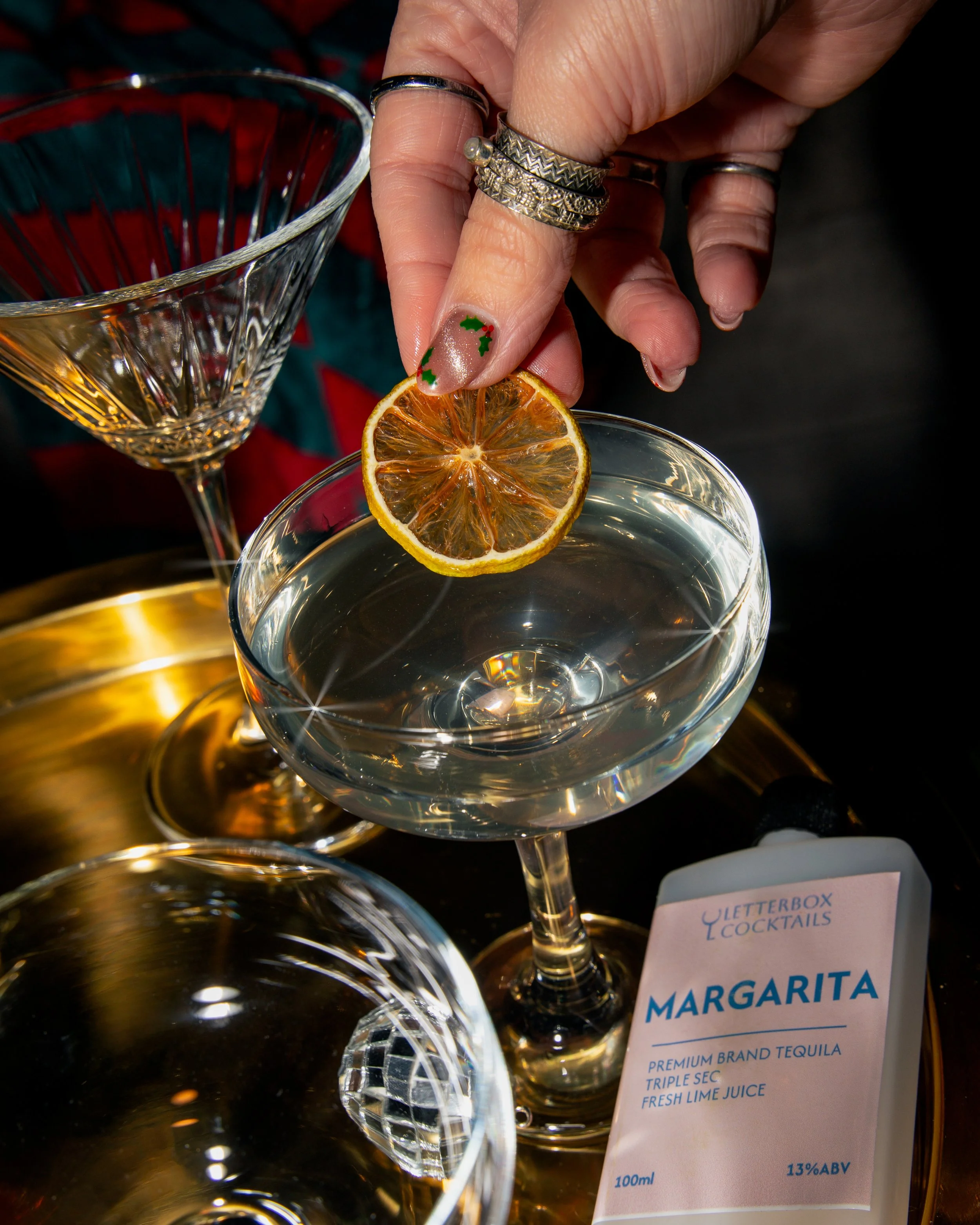 A hand garnishing a margarita with a dried lemon slice, surrounded by empty cocktail glasses and a bourbon labeled 'Margarita' with ingredients listed, on a gold tray.