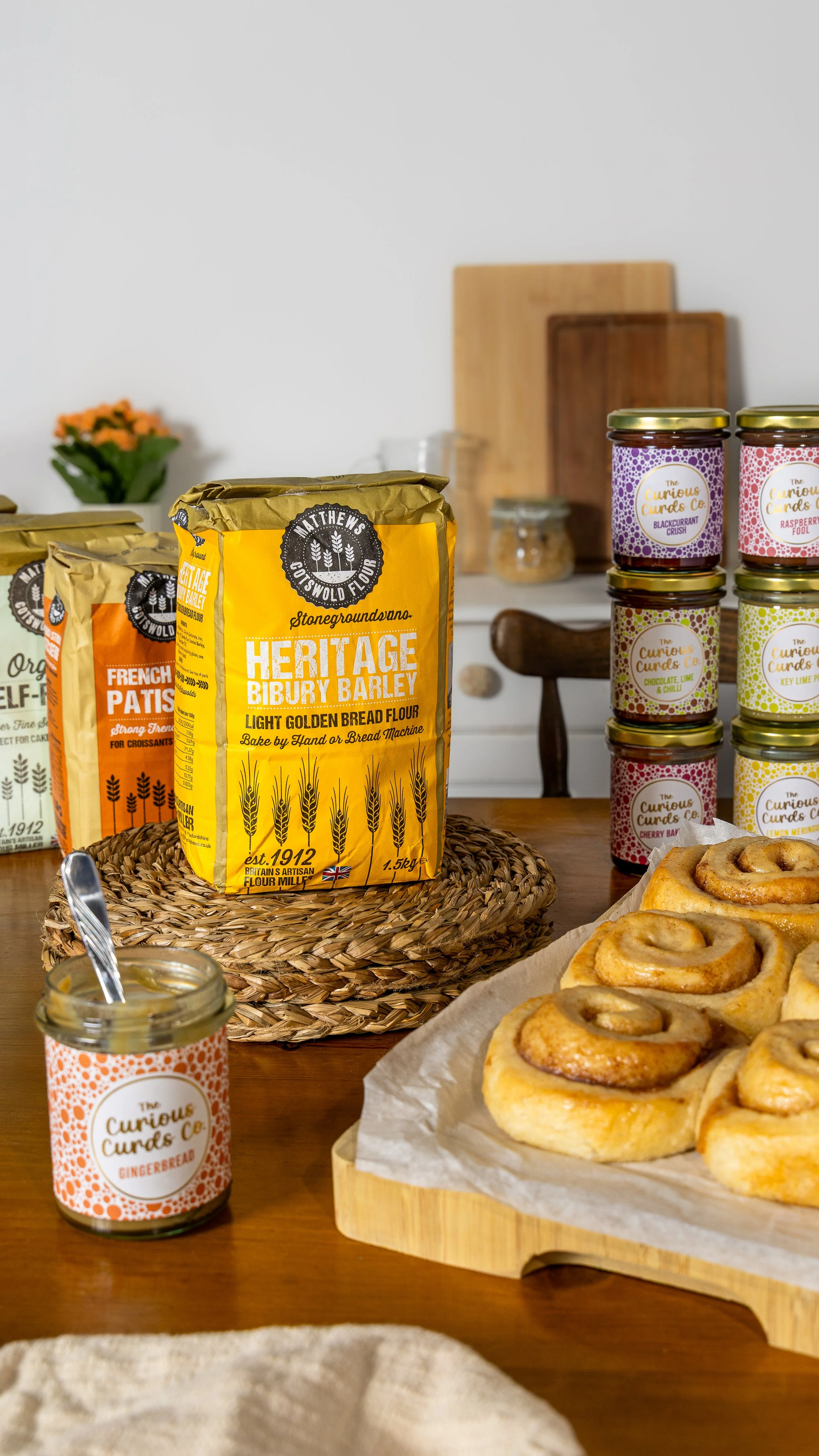 A bowl of cinnamon rolls on a wooden cutting board, a jar of gingerbread curd, a bag of heritage Bibury barley bread flour, and a stack of jars with colorful labels on a kitchen table.
