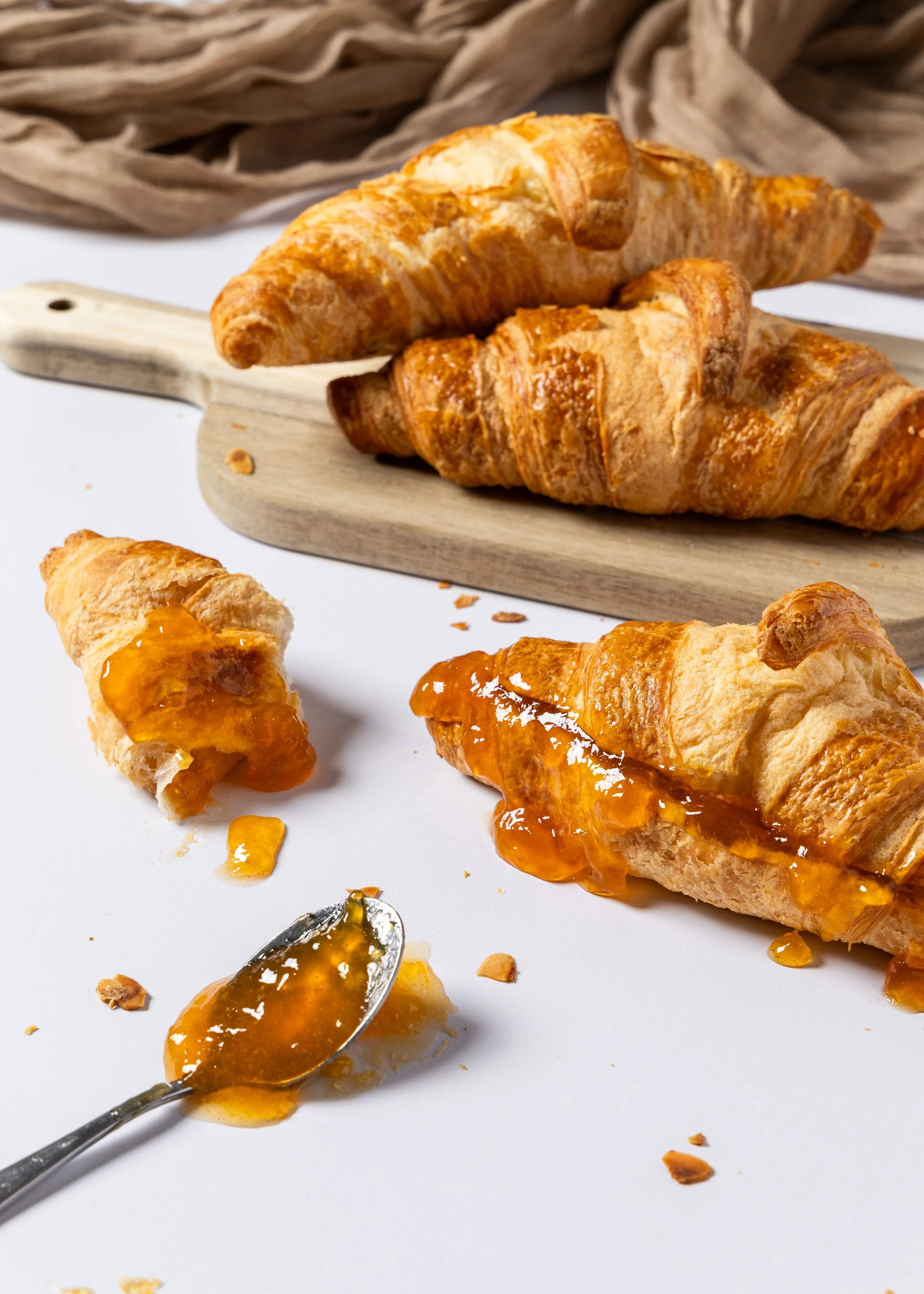 Two croissants, one with orange jam, on a white surface, with a wooden cutting board and more croissants in the background.