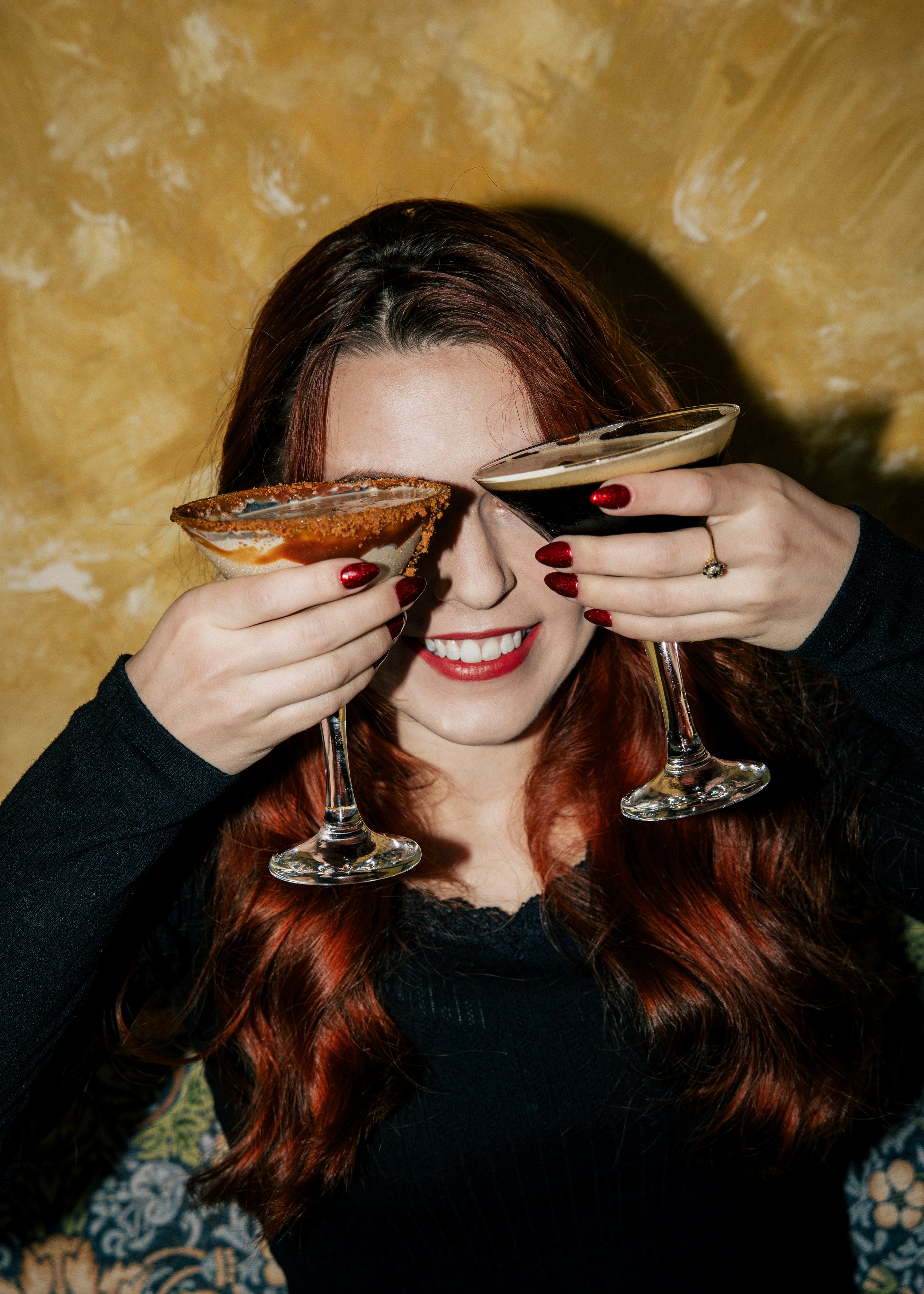 Woman with red hair smiling, holding two martini glasses with different drinks in front of her eyes, standing against a textured yellow wall.