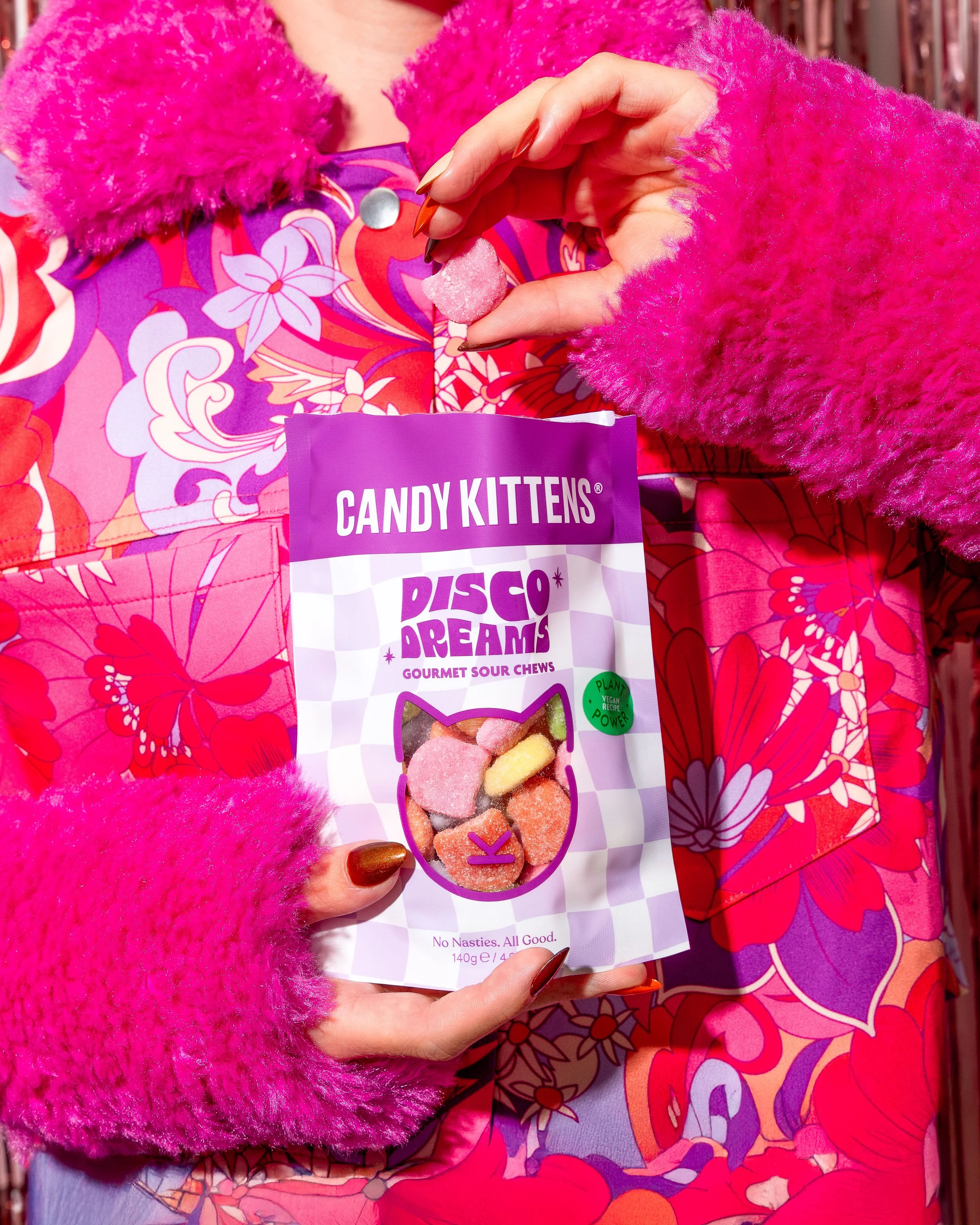 Close-up of a person holding a bag of Candy Kittens Disco Dreams gourmet sour chews, wearing bright pink fluffy gloves and a colorful floral jacket.