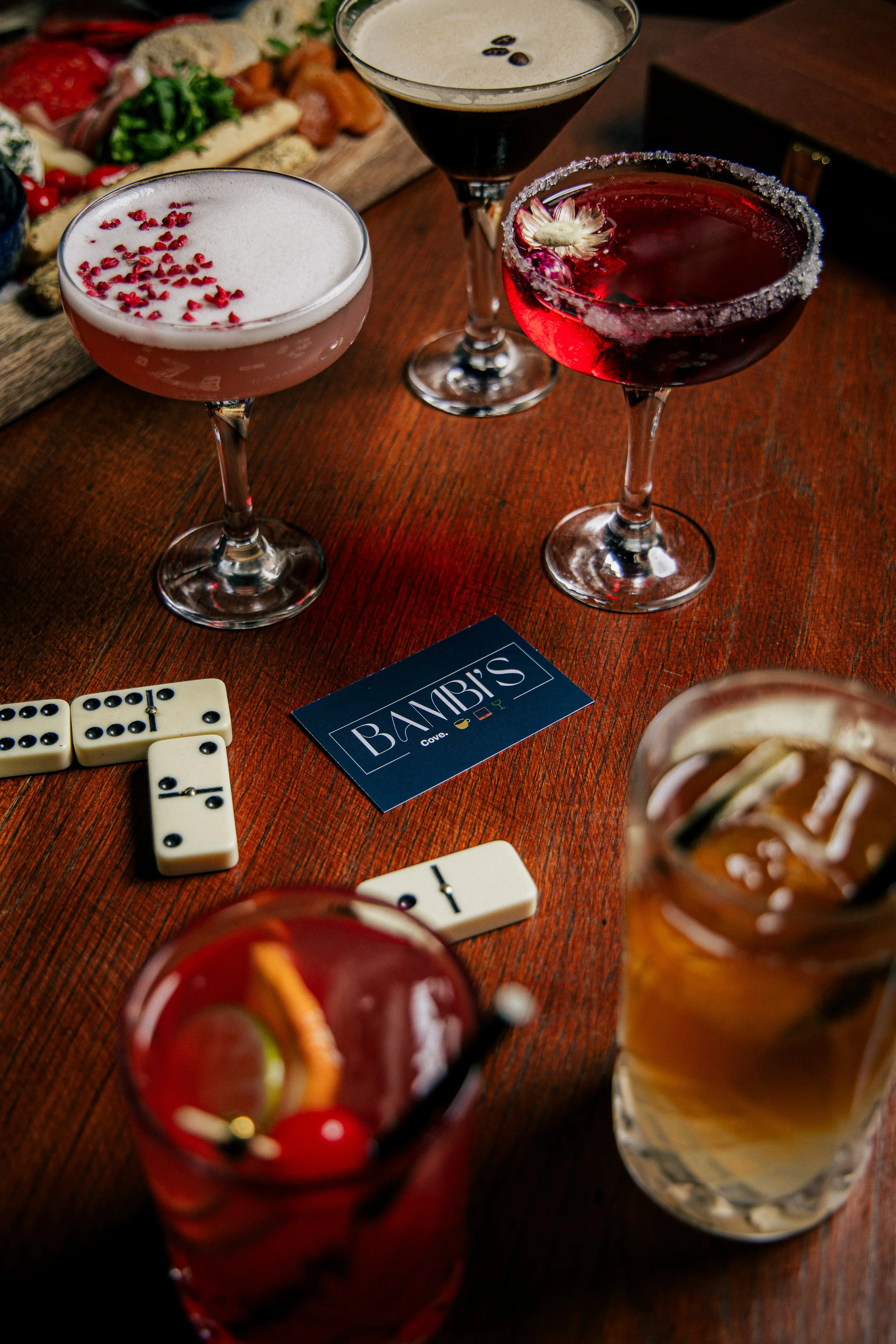 Three cocktails on a wooden table with a mixed drink in a jar, a dark martini with a flower garnish, and two other colorful cocktails, along with dominoes and a 