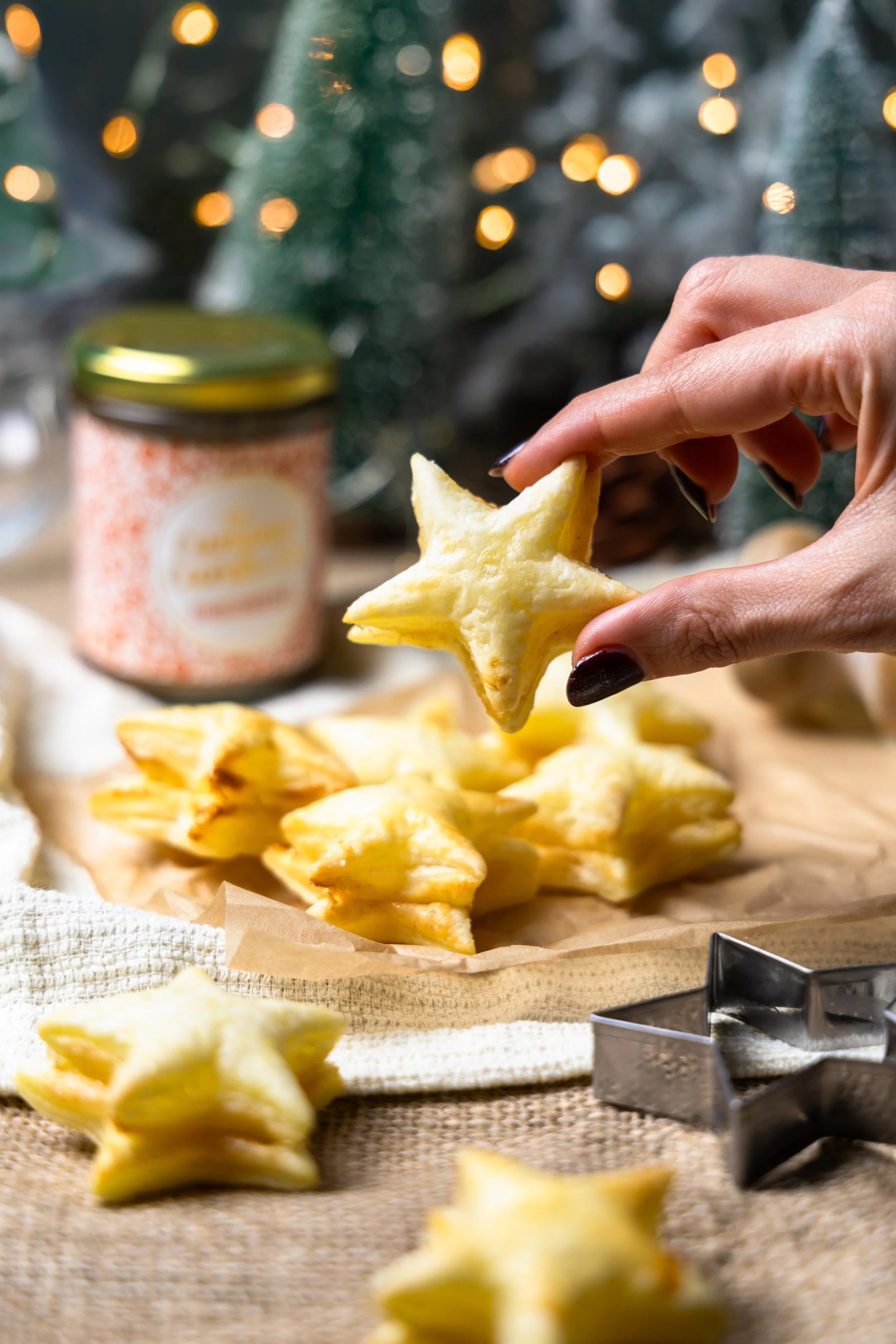 A hand holding a star-shaped cookie over a plate of similar cookies, with a festive background of Christmas lights and a jar on a table.