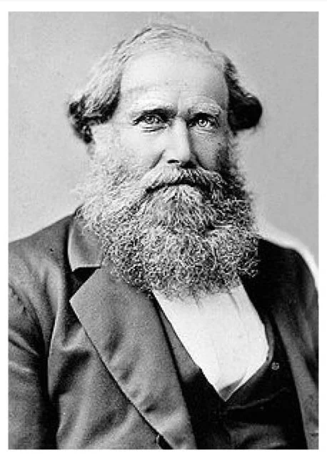 Black and white photograph of a man with a full beard and mustache, wearing a suit jacket and a collared shirt.
