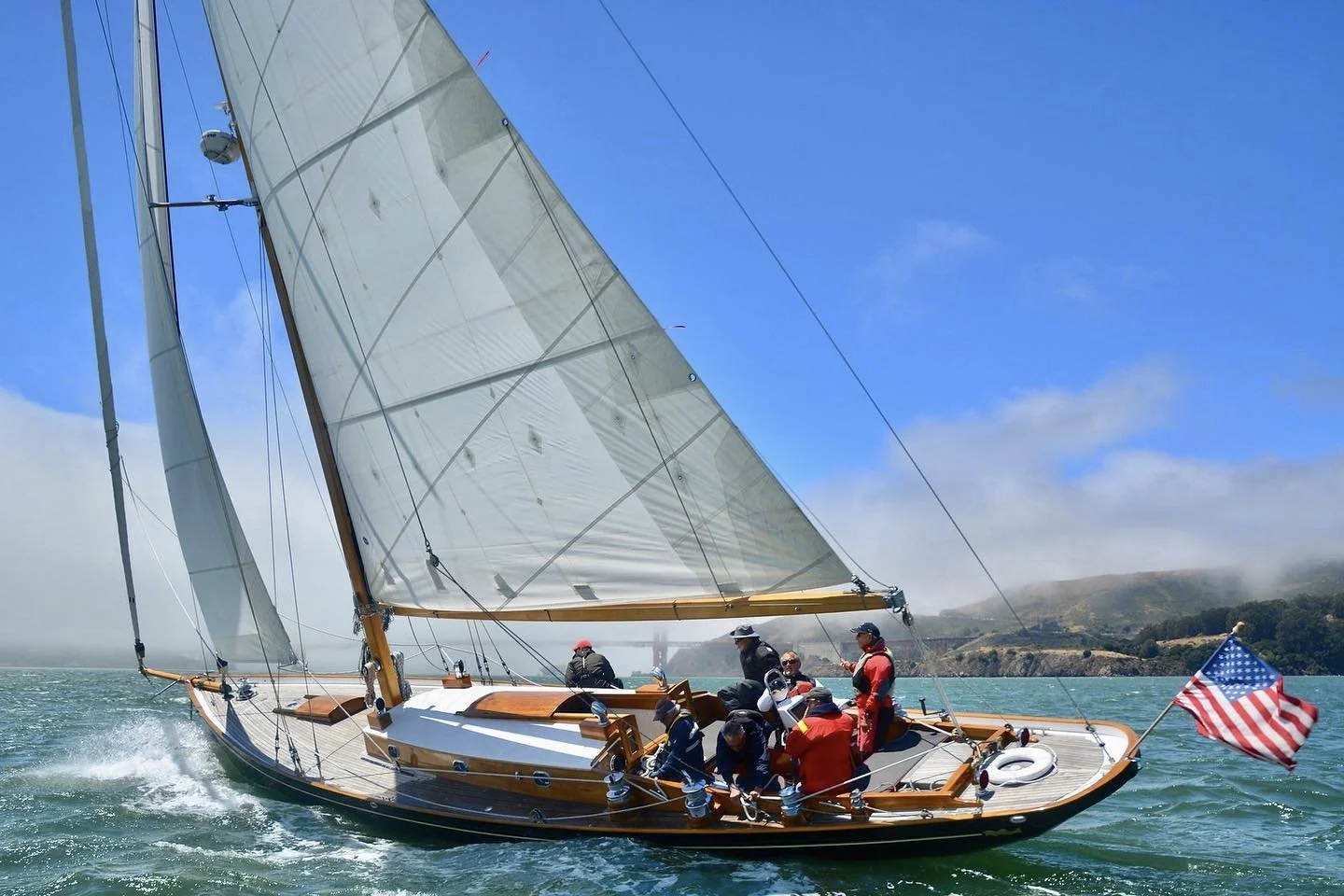 A sailboat with large tan sails on the water, carrying multiple people in life jackets and casual clothing, with an American flag at the stern and a scenic hilly coastline in the background under a partly cloudy sky.