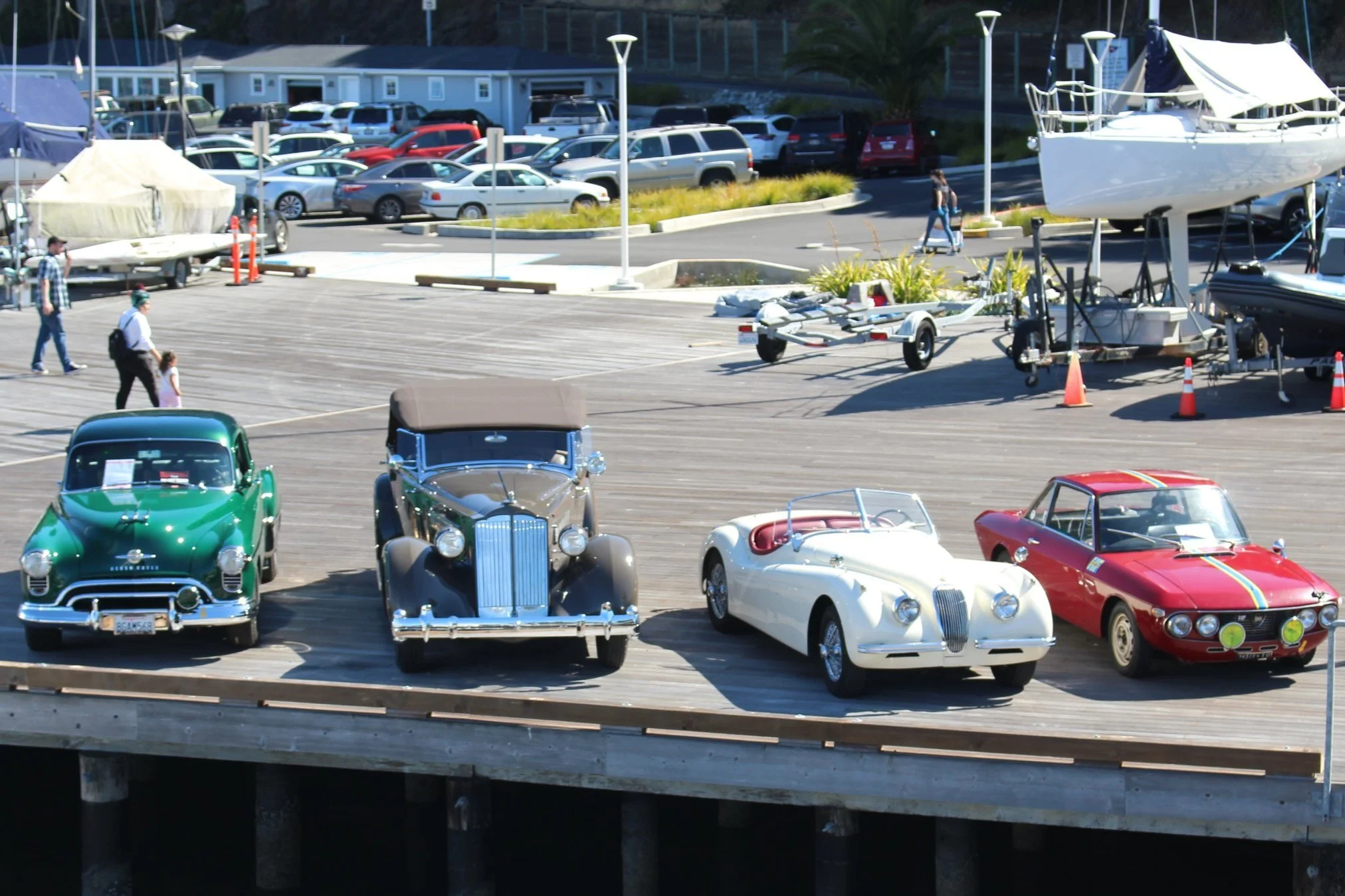 Four vintage cars parked on a wooden dock with boats and a parking lot in the background. From left to right, a green classic car, a silver vintage convertible, a white classic convertible, and a red racing car with yellow stripes.