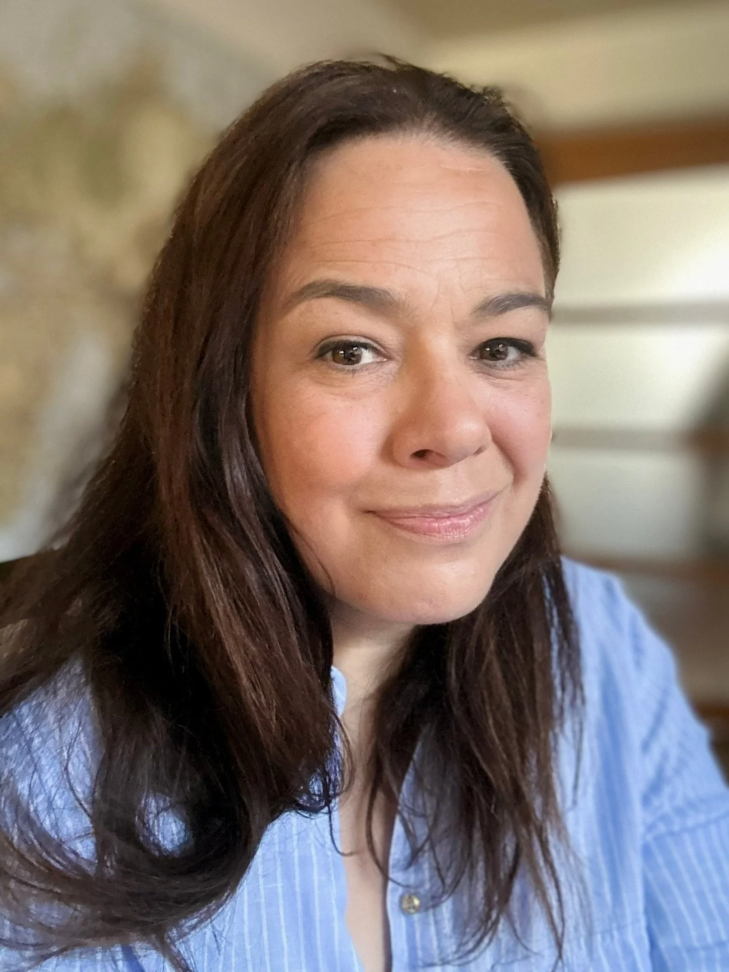 Close-up of a woman with long dark hair, wearing a blue shirt, smiling in a cozy indoor setting.