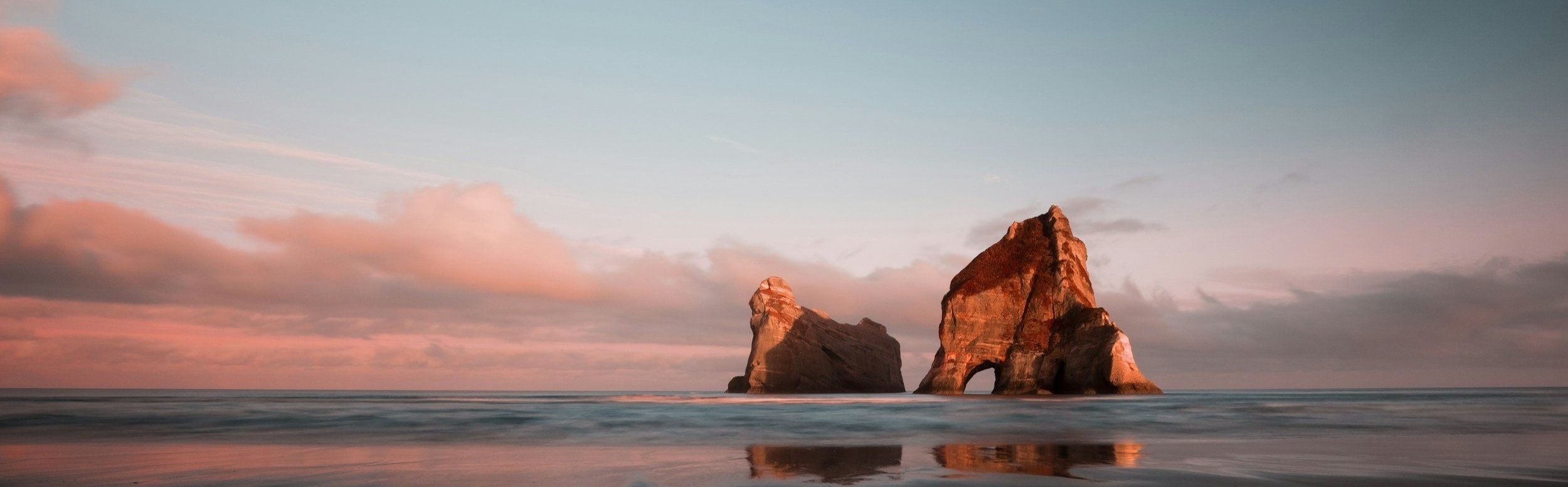 Two large rock formations in the ocean at sunset, with pink and orange clouds in the sky.