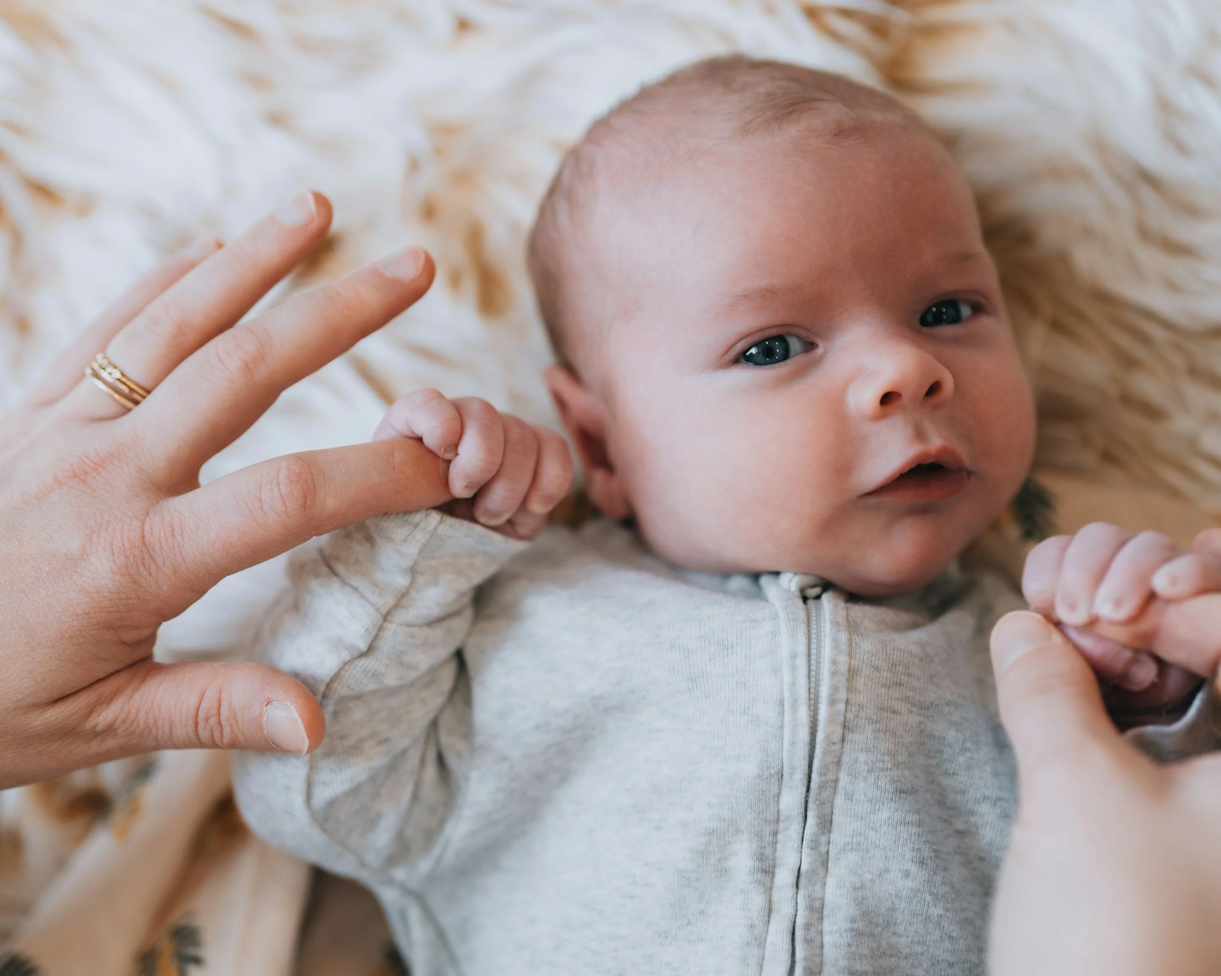 A baby lying on a soft, cream-colored blanket, holding an adult's fingers with both hands, looking at the camera.