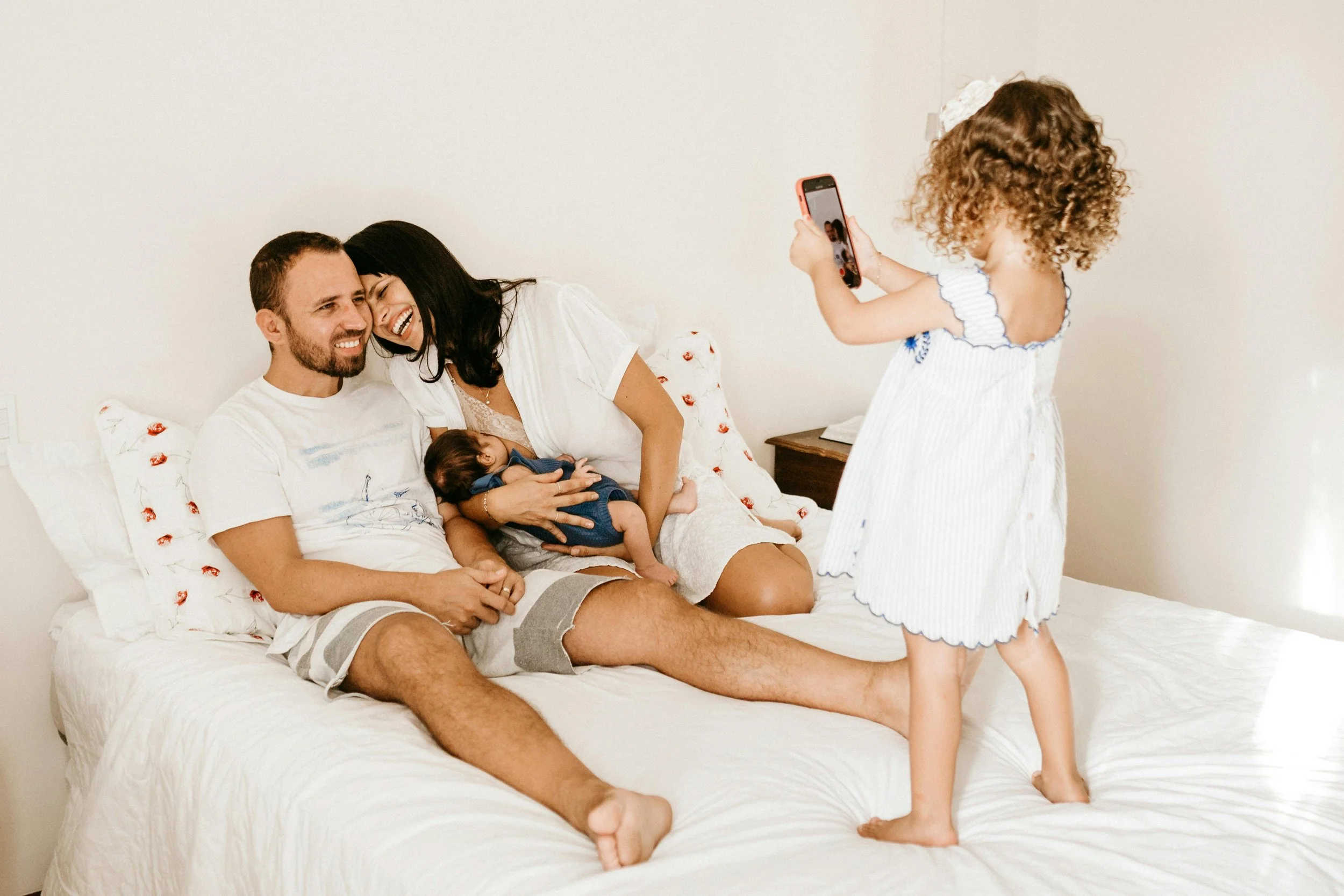 Family on bed taking a photo. Woman holds baby, man laughs, young girl in nightwear takes picture with phone.
