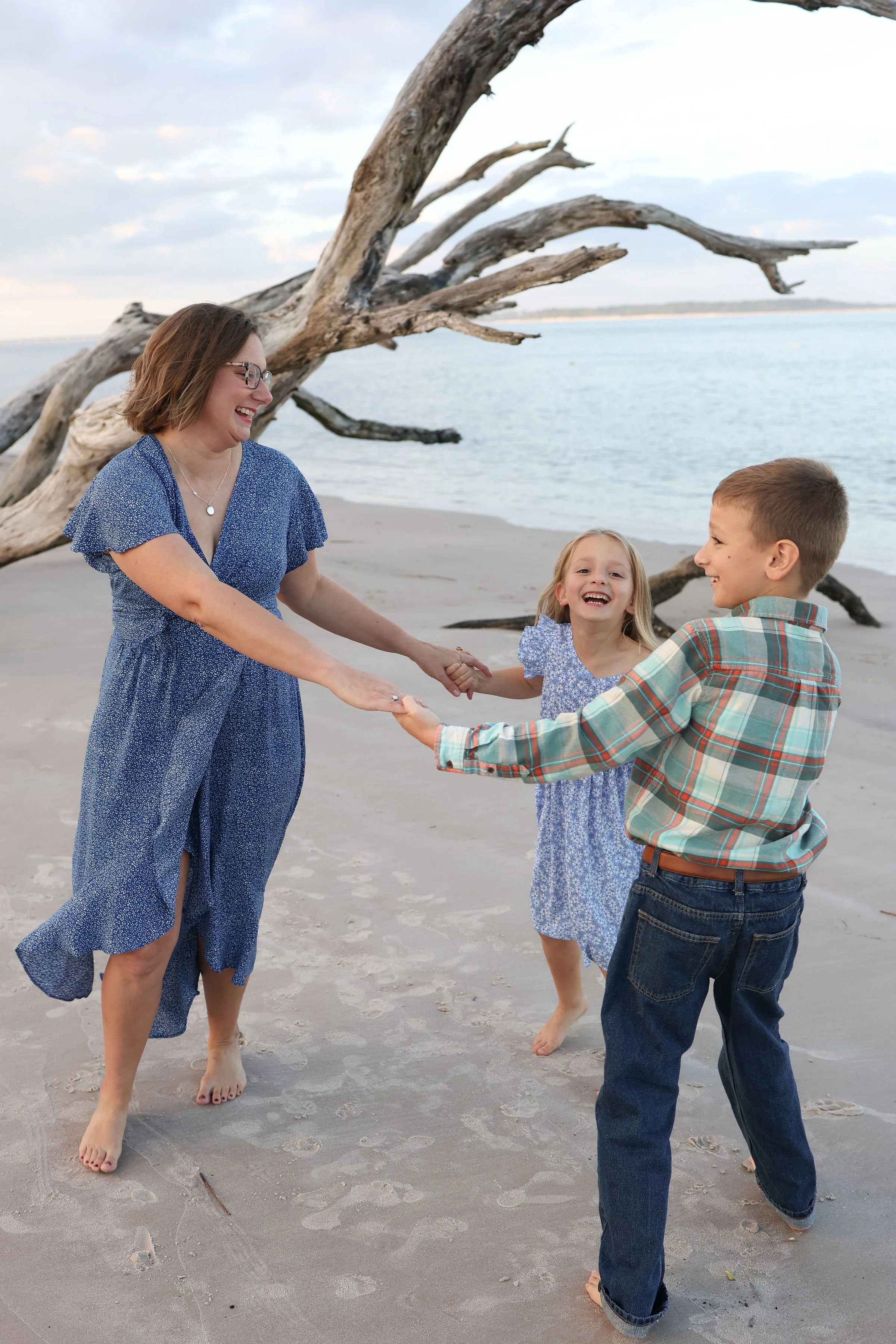 A woman and two children playing and laughing on a beach with a large driftwood tree in the background.