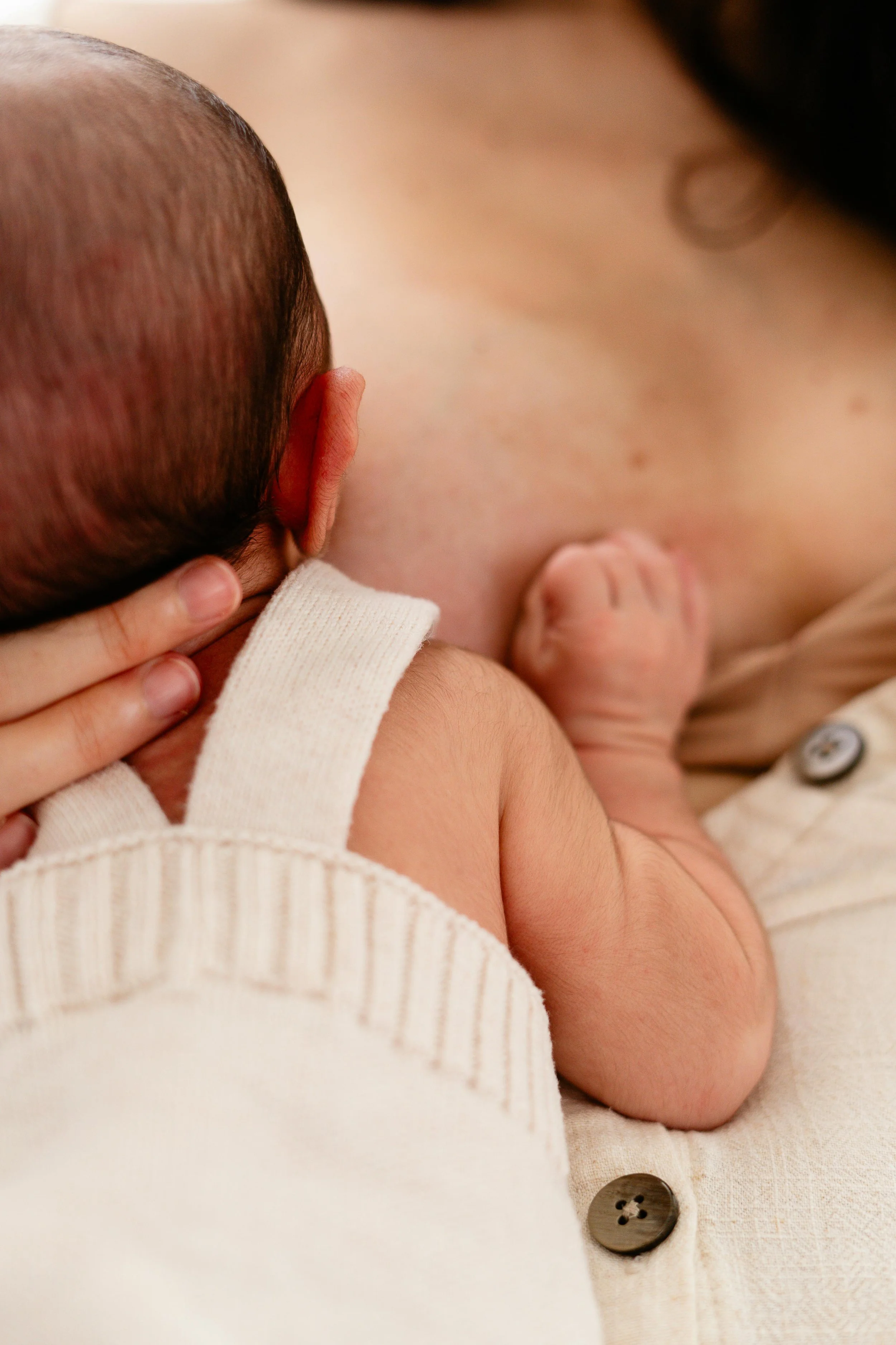 Close-up of a person holding a sleeping baby, showing the baby's head, ear, and tiny arm resting on the person's chest. The baby is wearing a cream-colored outfit with a button.