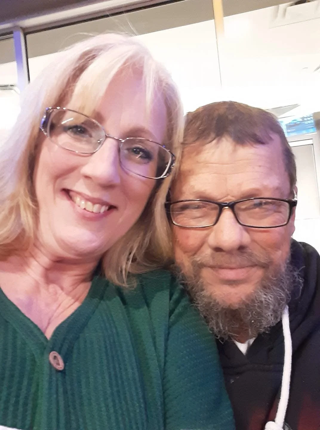 A smiling woman with blonde hair and glasses, and a smiling man with glasses and a beard, taking a close-up selfie together.