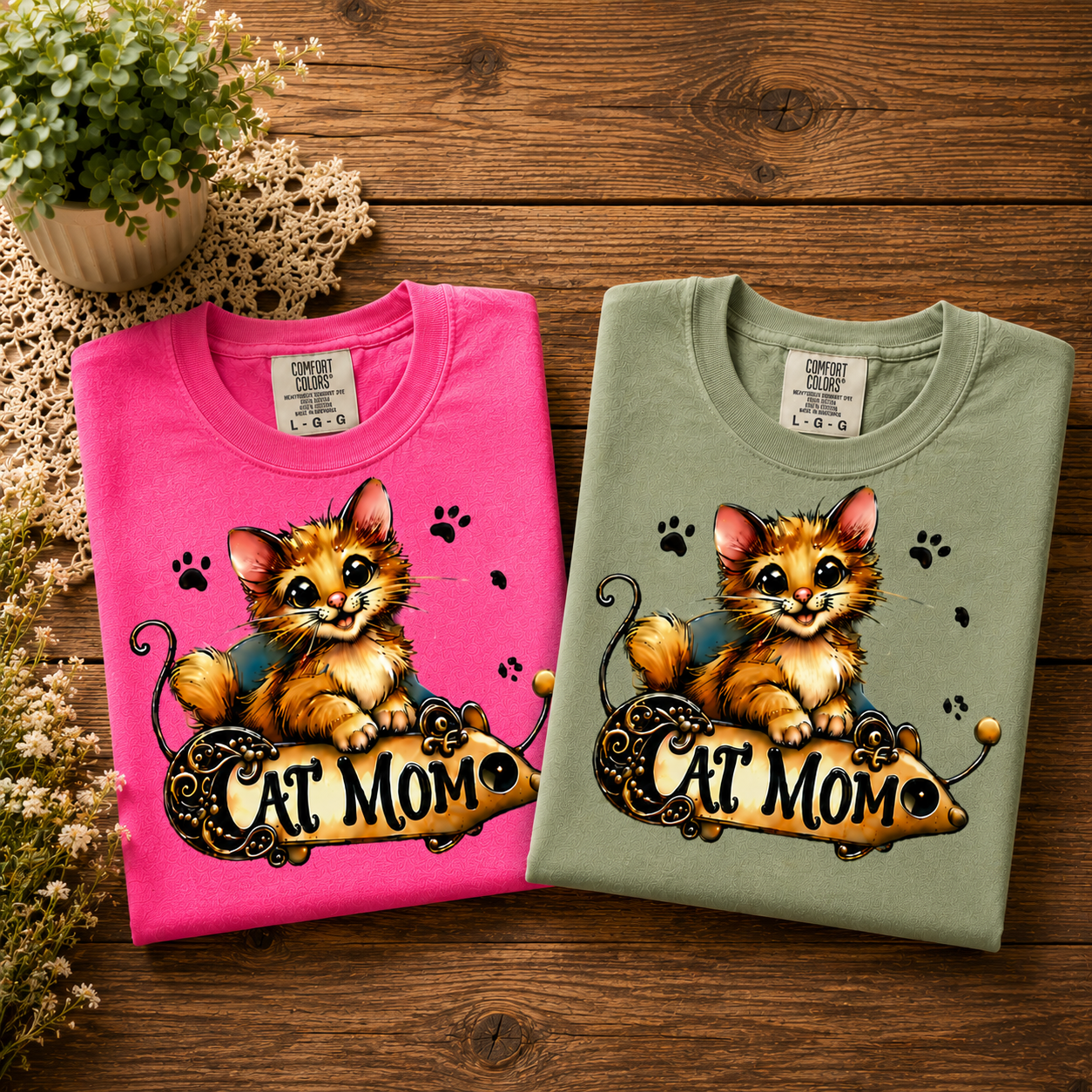 Two folded t-shirts, one pink and one light green, featuring a cute cartoon kitten with large eyes and a playful expression, sitting on a decorative banner that reads "Cat Mom." The shirts are placed on a wooden surface with a small potted plant and some flowers nearby.