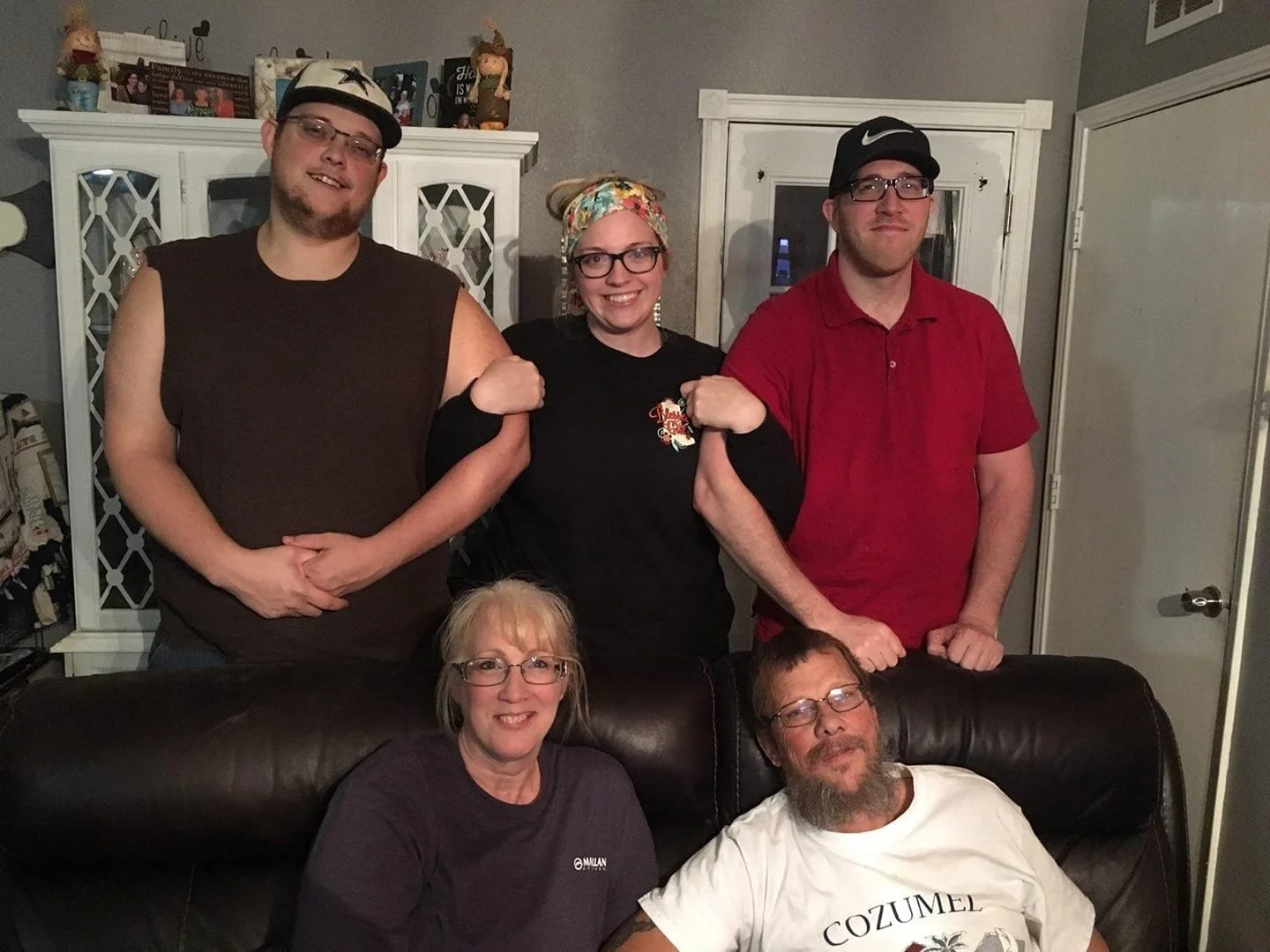 A group of six people, three men and three women, are gathered in a living room, posing for a photo. Some are sitting on a sofa, while others are standing behind it. There is a white cabinet with decorative glass doors and various ornaments on top in the background.