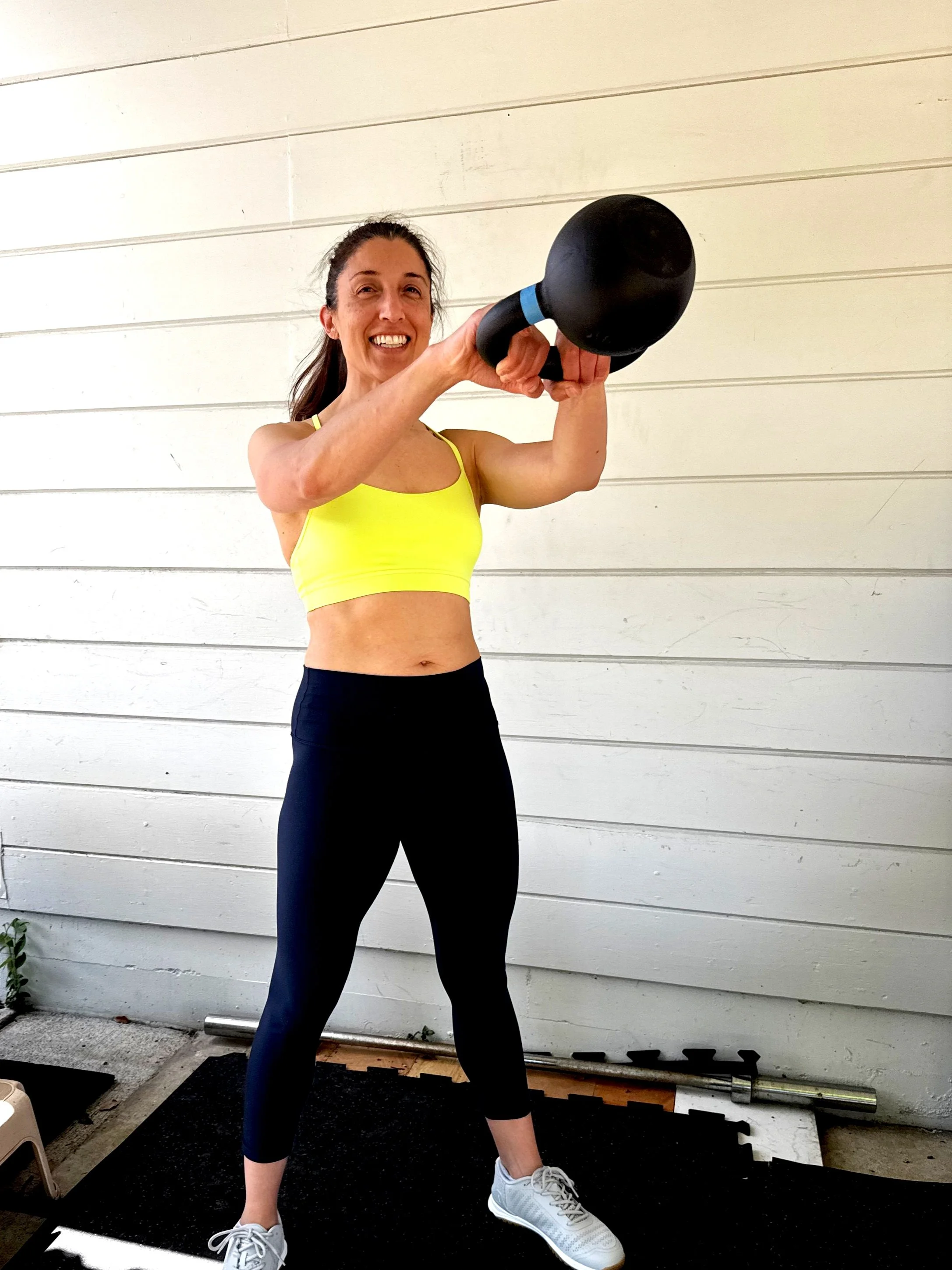 A smiling woman in yellow activewear swinging a kettlebell.