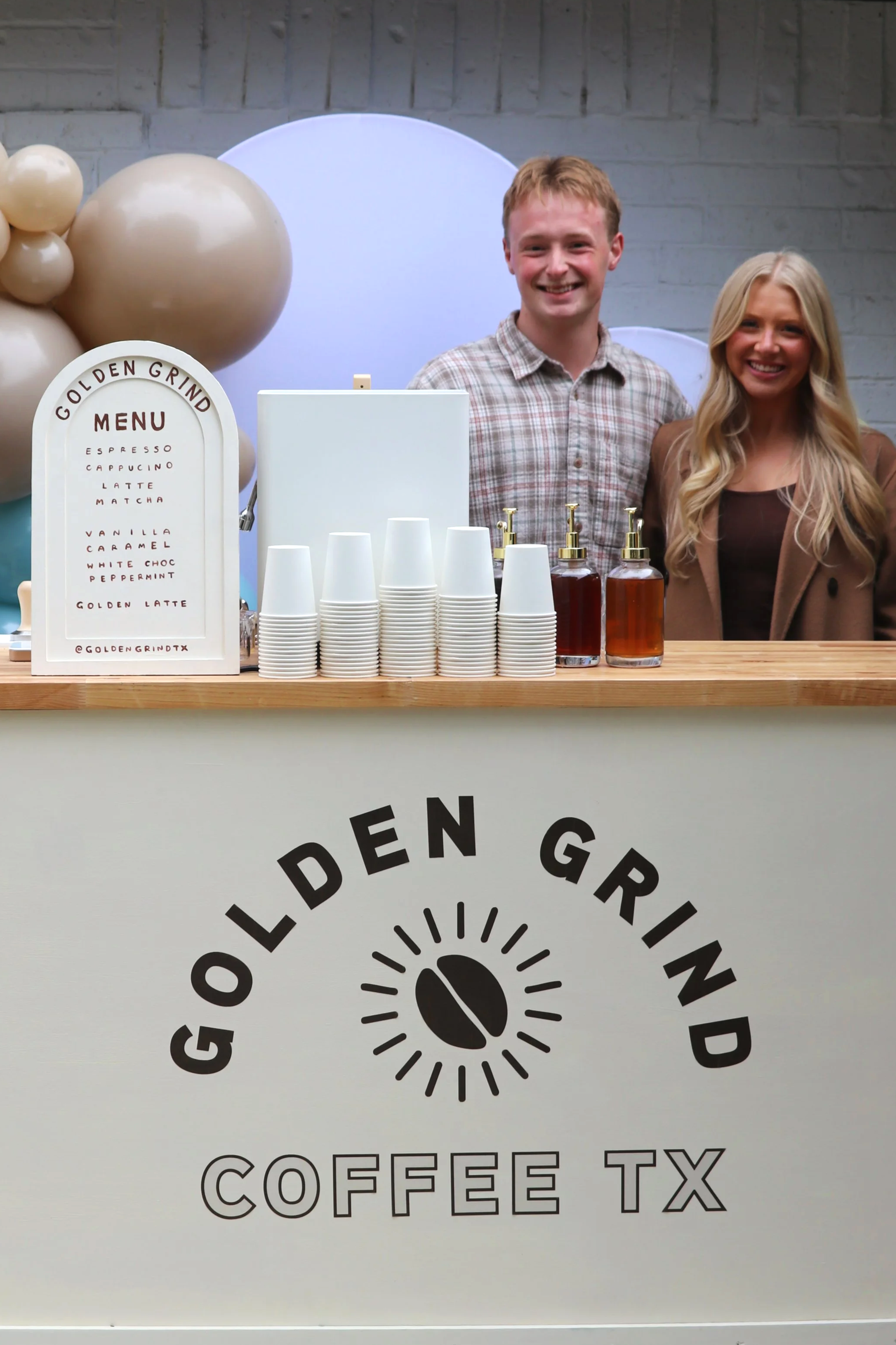 Smiling man and woman serving coffee at Golden Grind Coffee stand, with cups, syrup bottles, and menu on display.