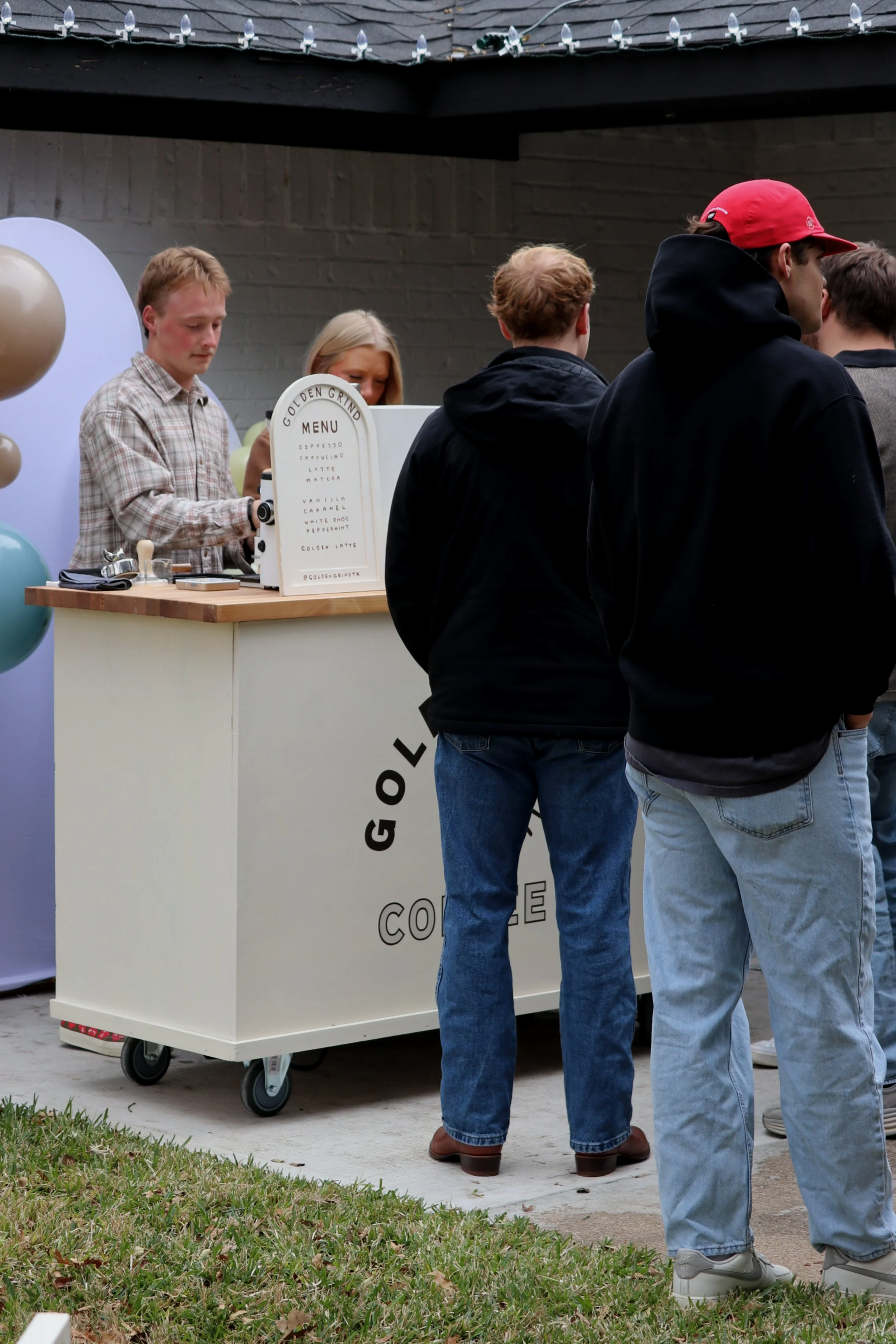 People waiting in line at a coffee stand, with a staff member serving coffee behind the counter decorated with balloons.