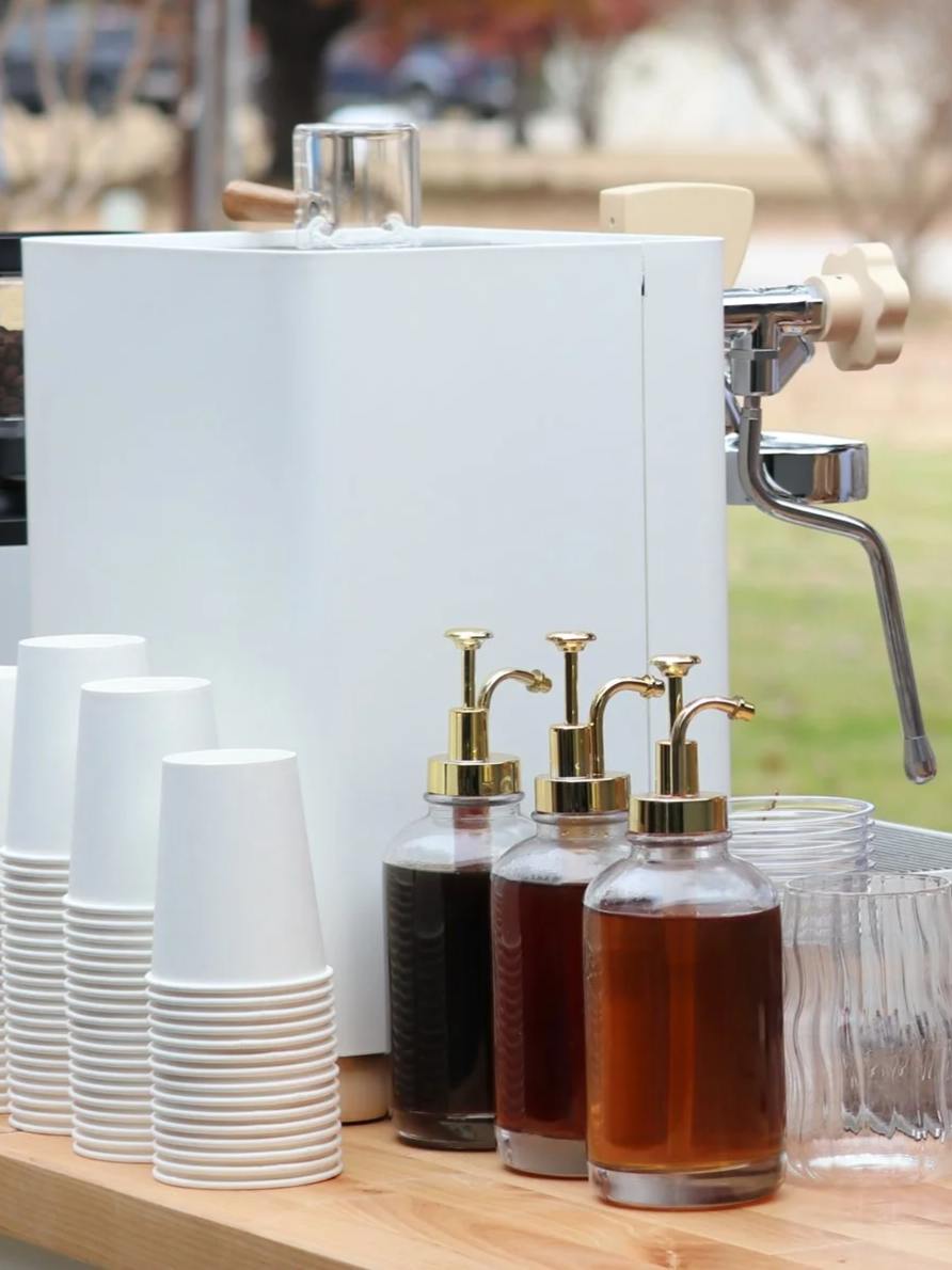 Outdoor coffee station with white paper cups, glass syrup bottles with pumps, and a white coffee machine.