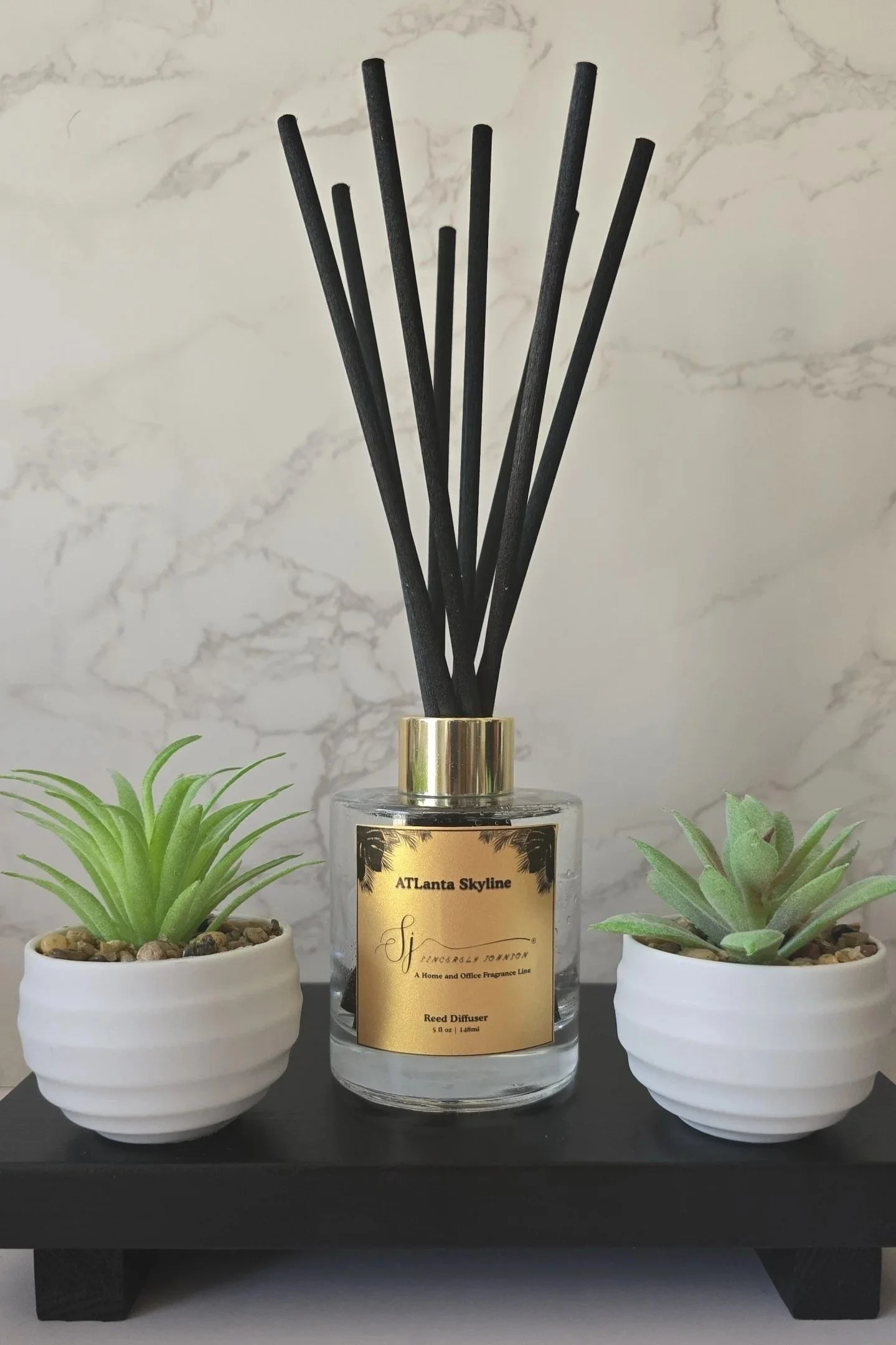 A reed diffuser with black sticks in a clear glass bottle labeled "ATLanta Skyline" is placed on a black wooden platform, flanked by two small potted succulents in white ceramic pots.