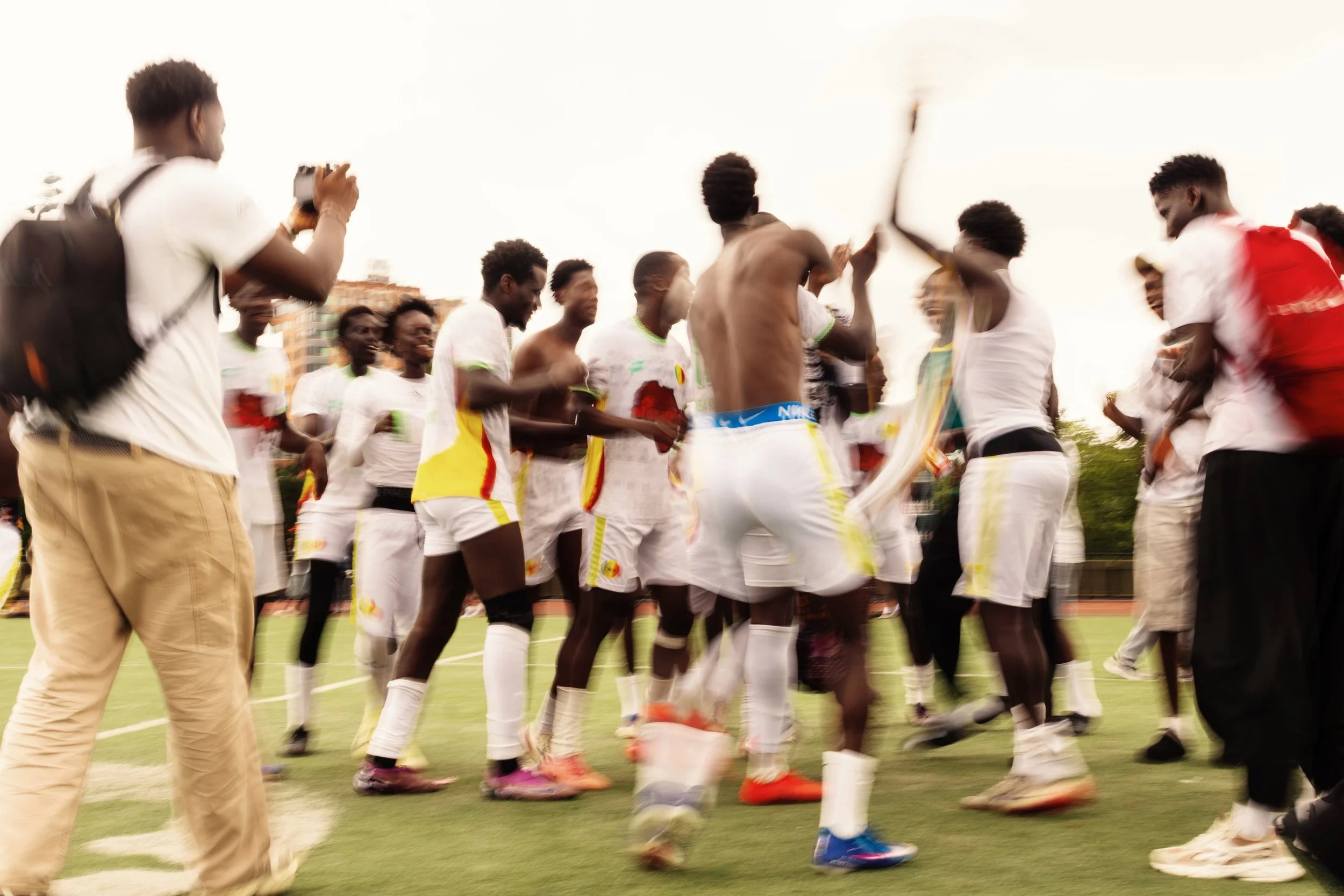 A group of people on a sports field, some in soccer uniforms, appear to be celebrating or playing a game, with individuals smiling and engaging with each other.