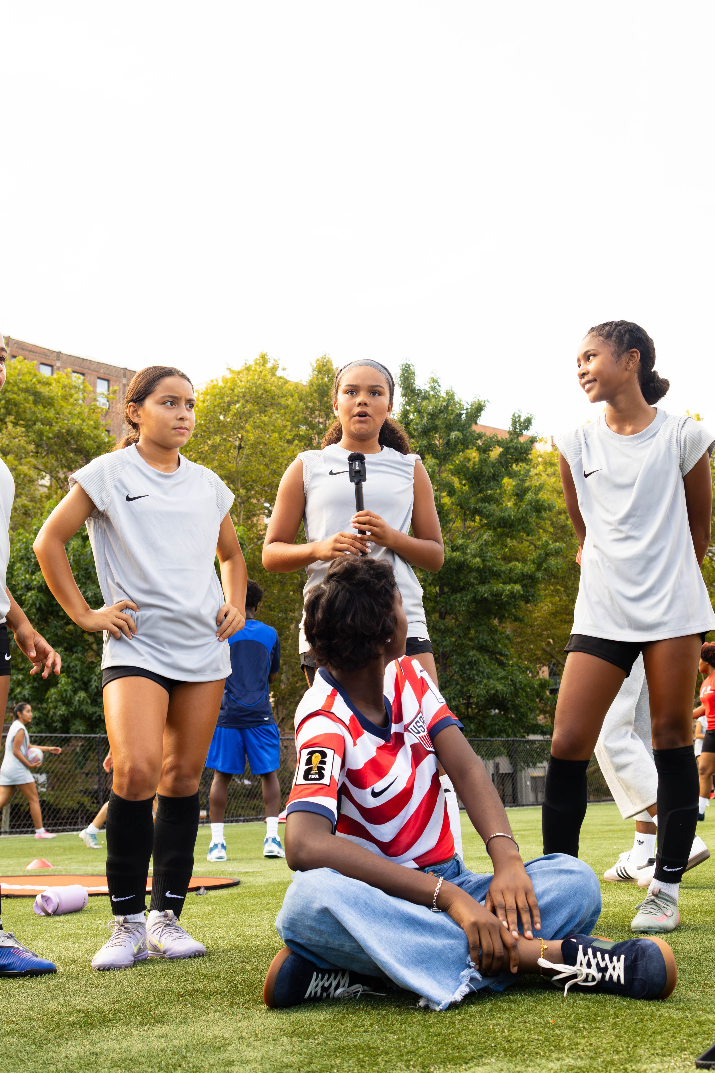 A group of young girls, some standing and one sitting cross-legged on the grass, at an outdoor sports field with green trees and buildings in the background. The girls are wearing athletic clothing, including soccer jerseys and shorts.