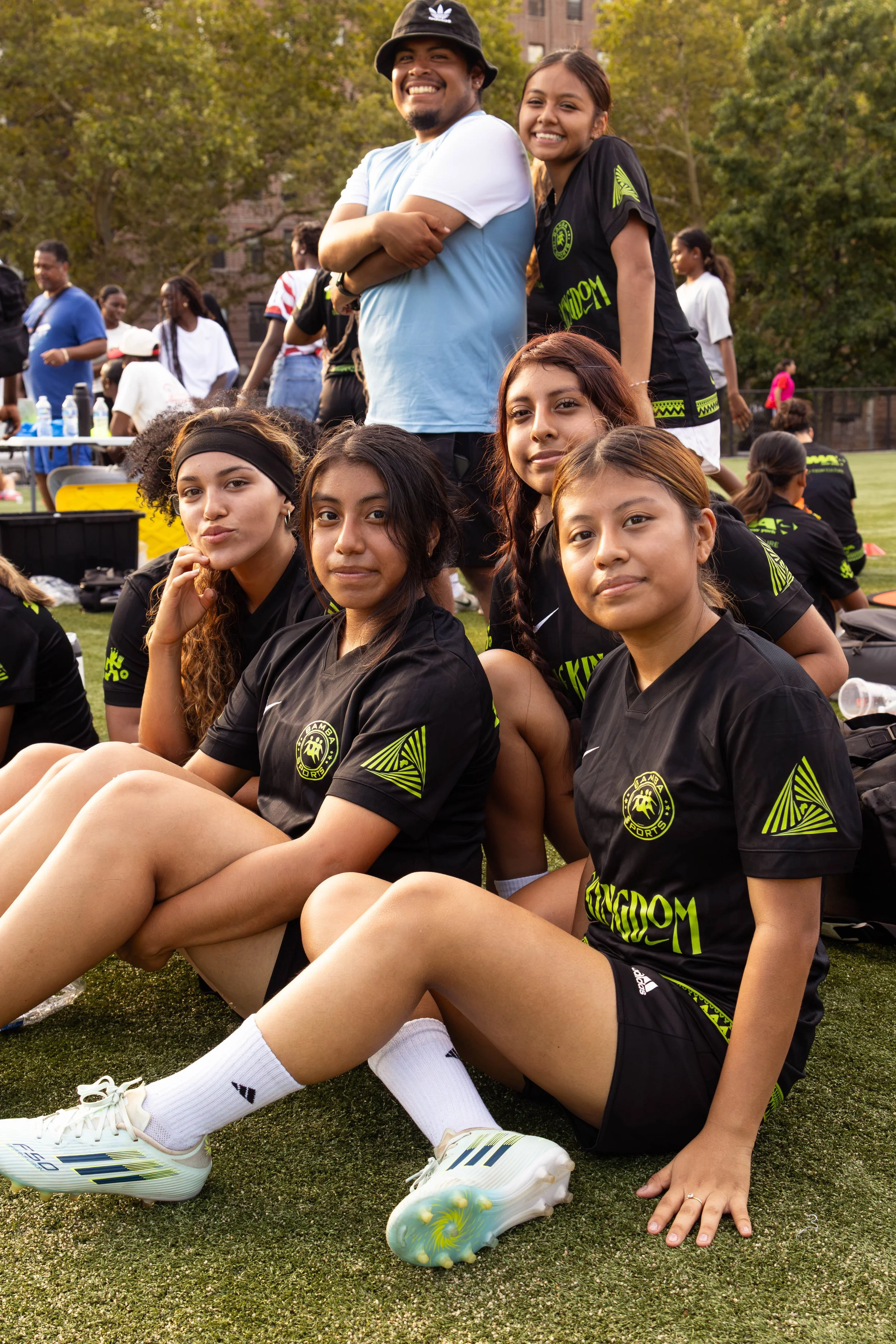 Group of young girls in black sports uniforms sitting on the grass at a sports event, with some smiling at the camera and others looking away, in an outdoor setting with trees and people in the background.