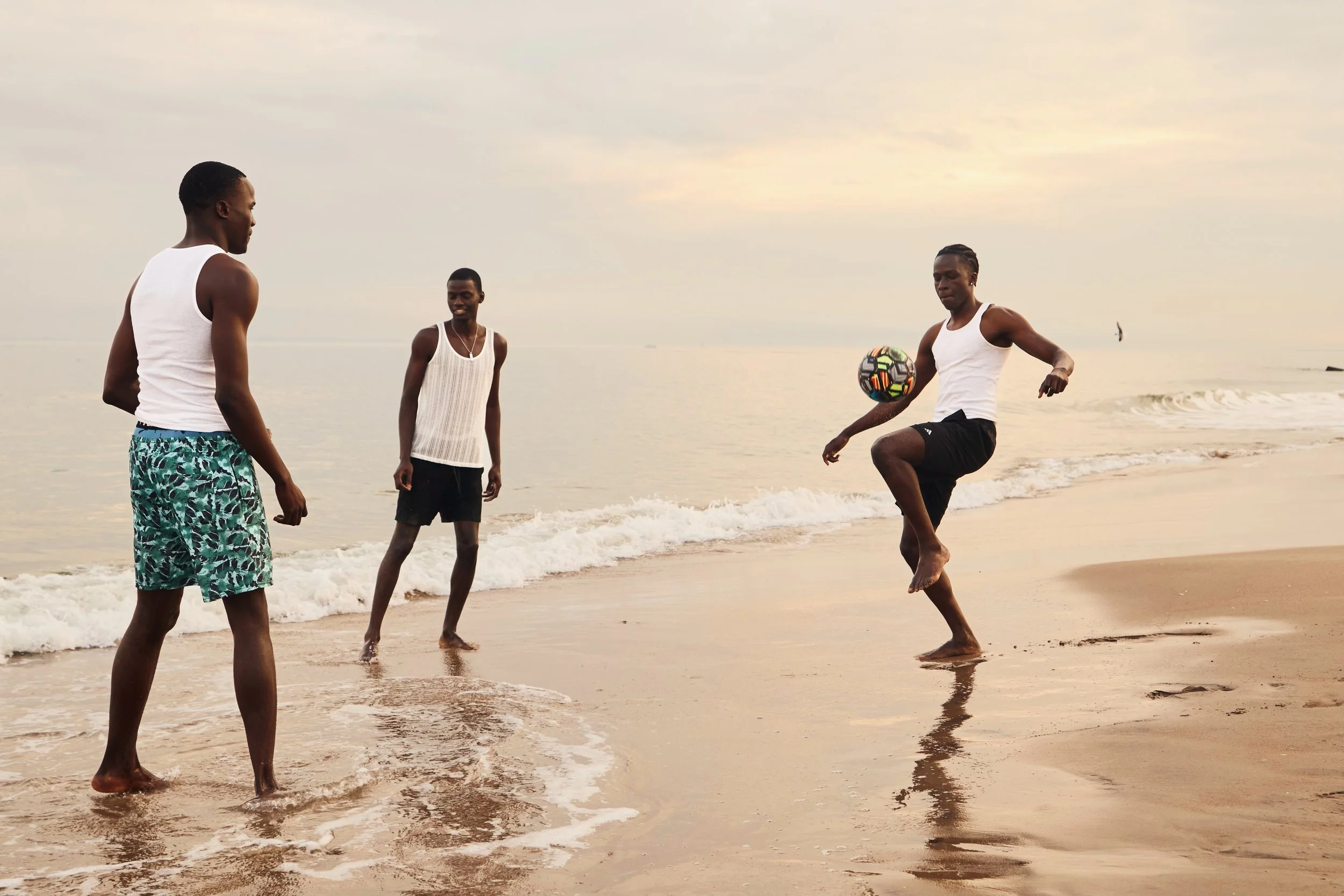 Four men playing soccer on a beach during sunset or sunrise with calm water and a sailboat in the background.