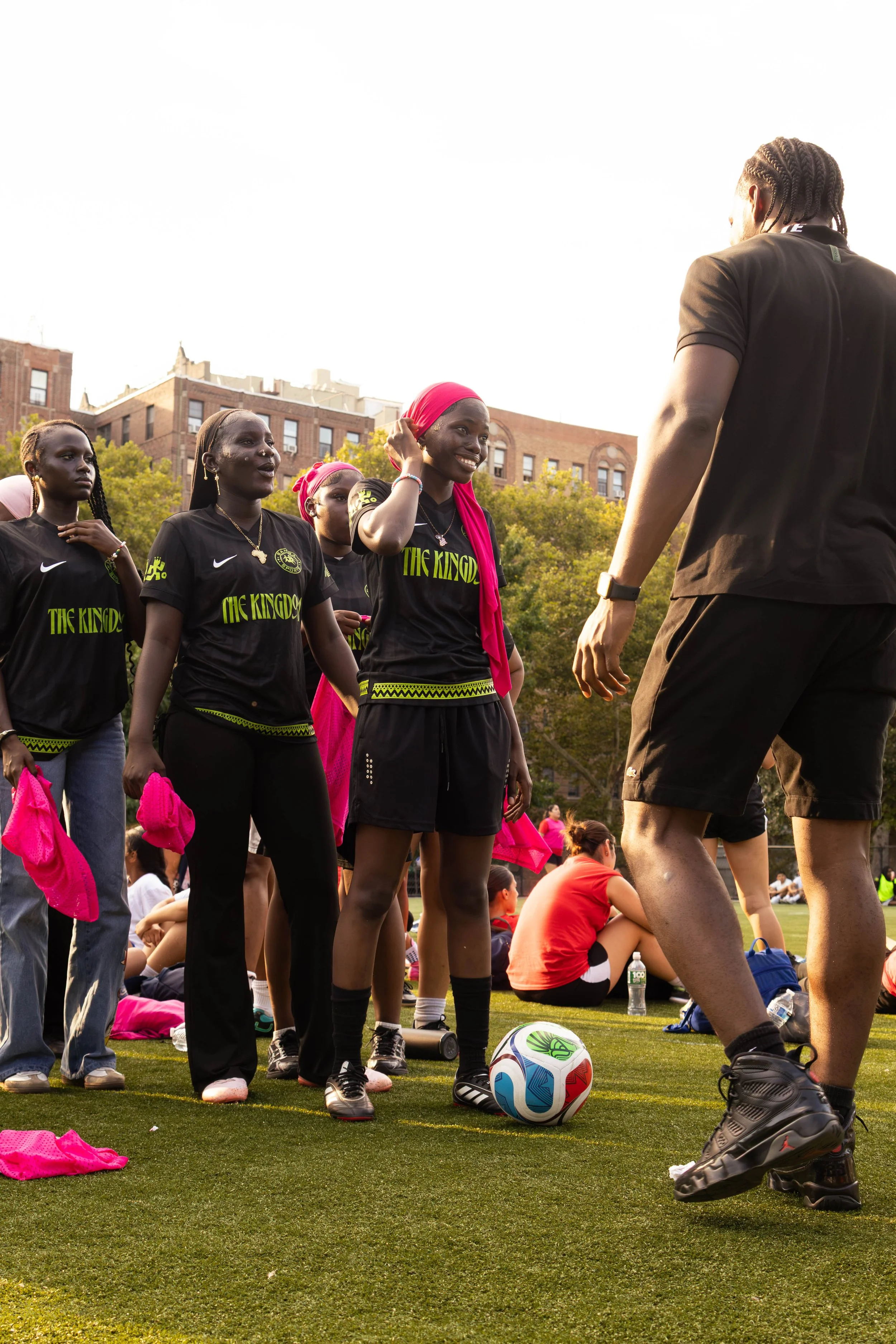 A group of women and a man gathered on a soccer field, some wearing black shirts with 'The Kingdom' written on them, with a soccer ball at their feet and other people sitting in the background, trees, and brick buildings.