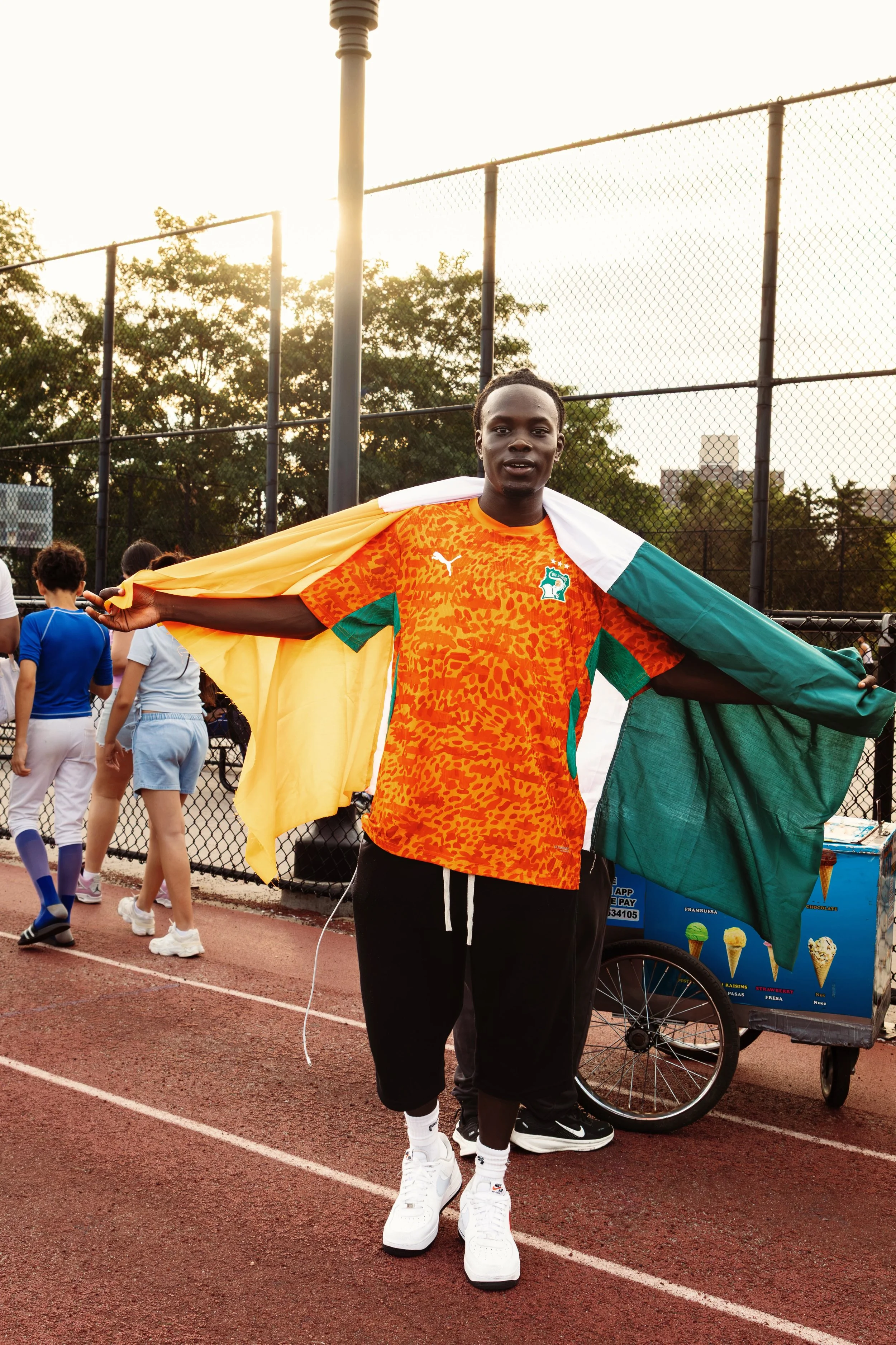 A young man on a track field holding a flag over his shoulders, wearing a colorful sports shirt and sneakers, with people and trees in the background during sunset.