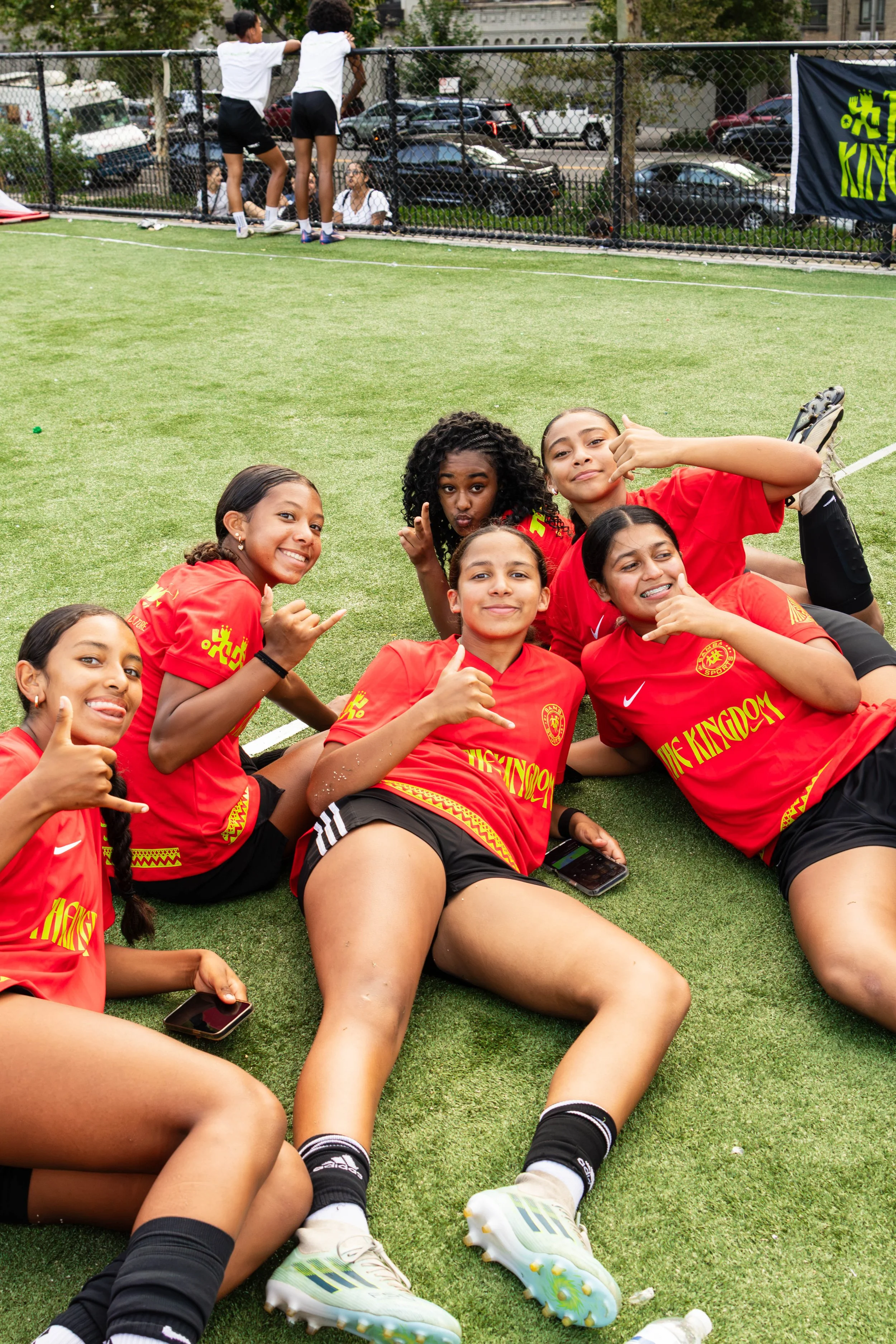 Group of young female soccer players in red jerseys lying on the grass field, smiling and giving thumbs-up, with some holding smartphones, during a break or after a game.
