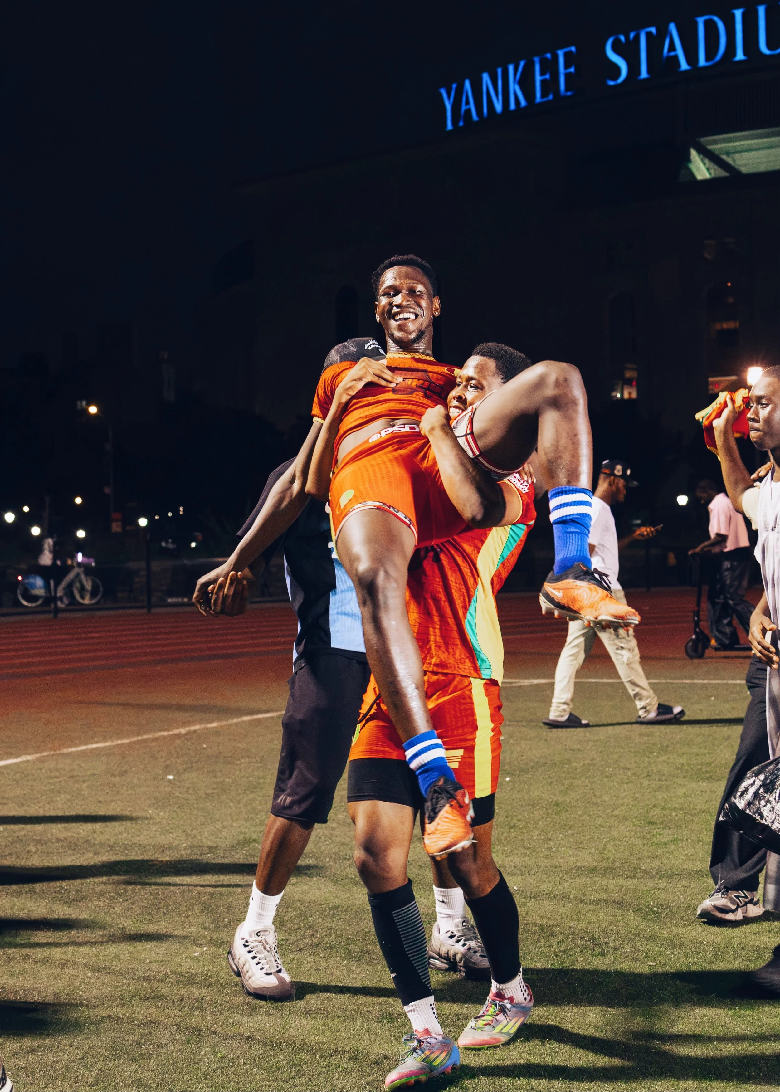 Celebrating athletes at Yankee Stadium; two men in sports attire being lifted and hugging after a sporting event at night.