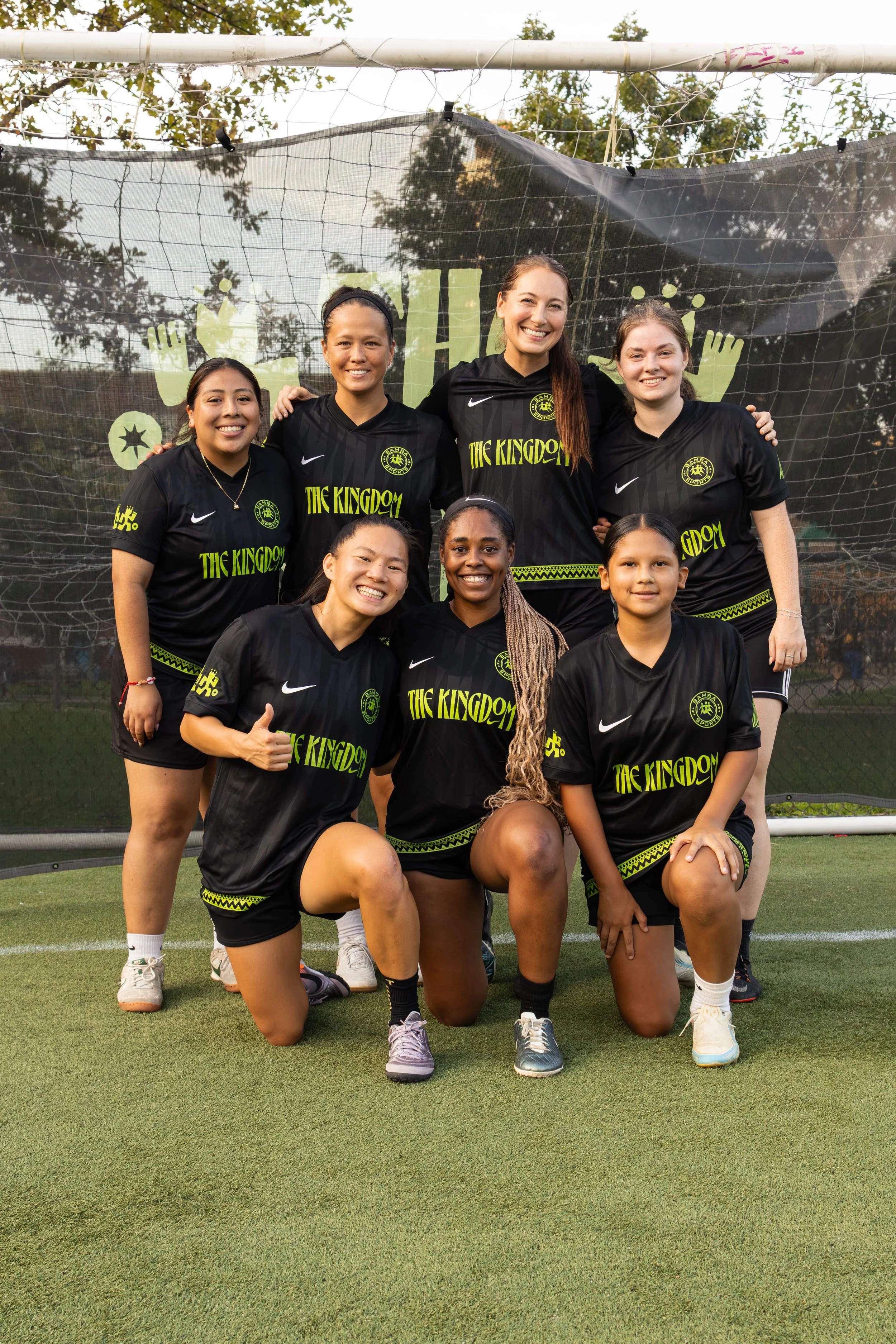 Group of seven women soccer players in black uniforms with the words "The Kingdom" on the front, posing on a soccer field in front of a goal net and black banner with lime green graphics, trees in the background.