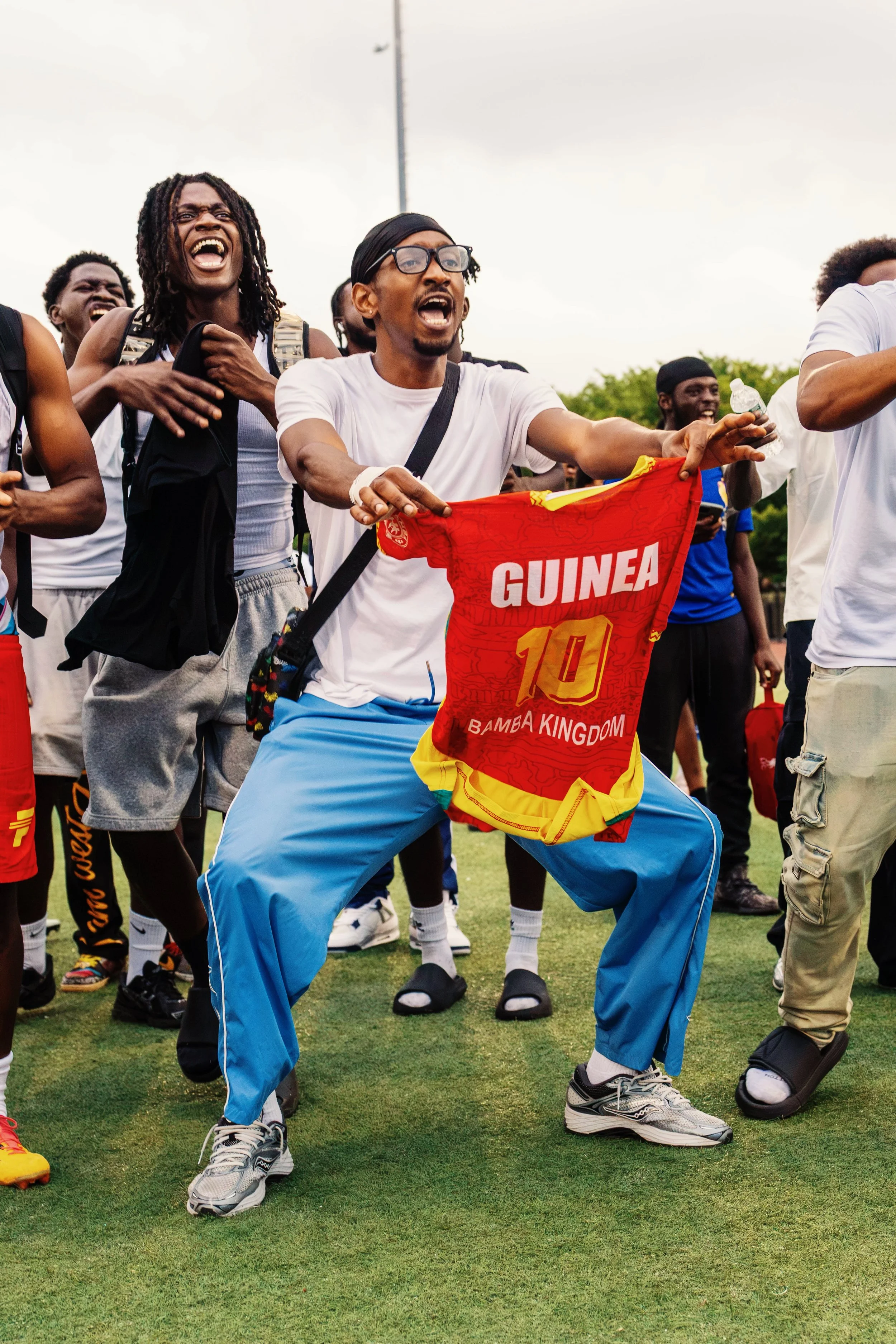 A group of people, some smiling and some shouting, standing on a grassy field. One person in the foreground holds a red and yellow flag that says "GUINEA" and wears blue pants, a white shirt, and gray sneakers.