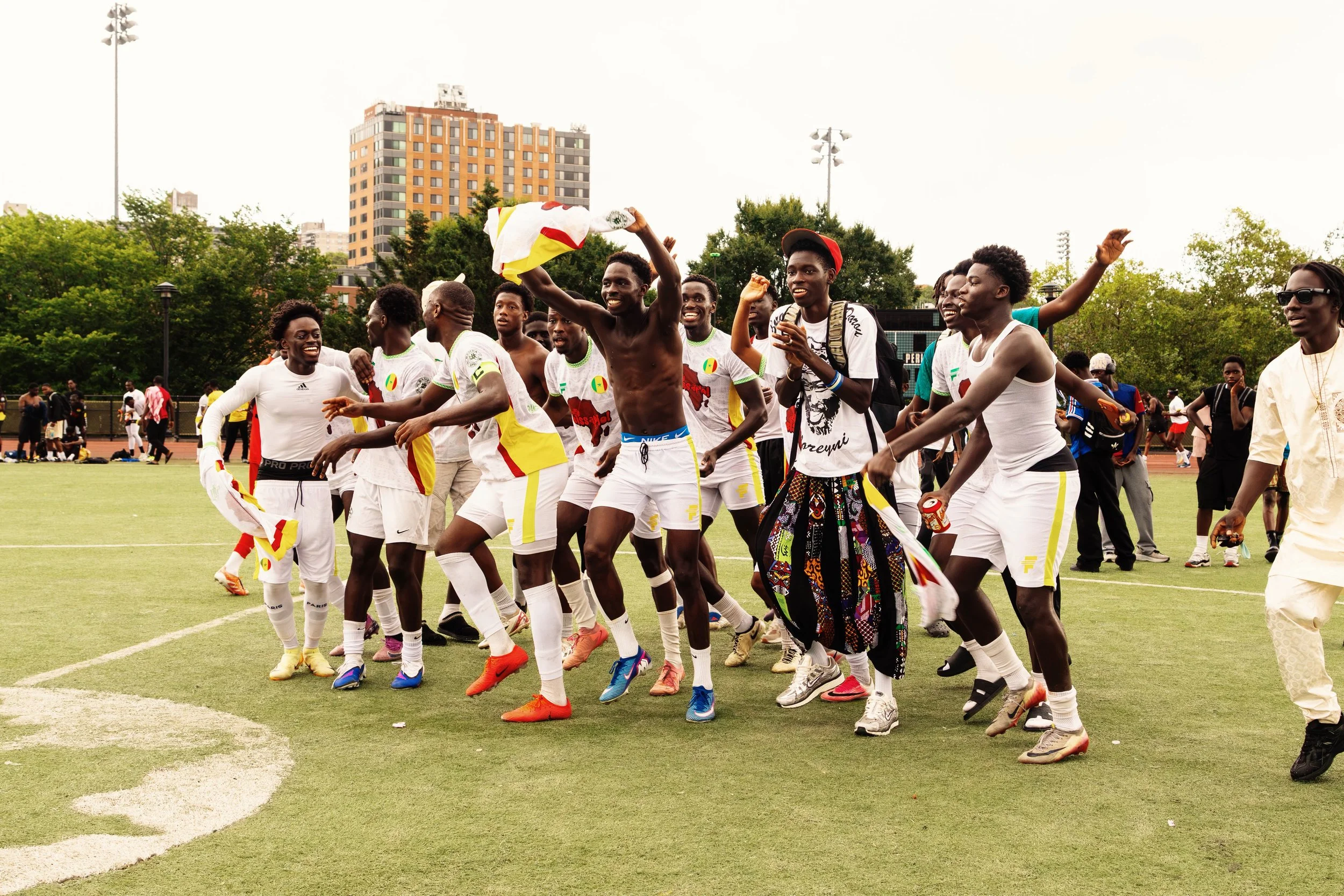 A group of celebratory soccer players on a field, some smiling, some cheering, with trees and a building in the background.